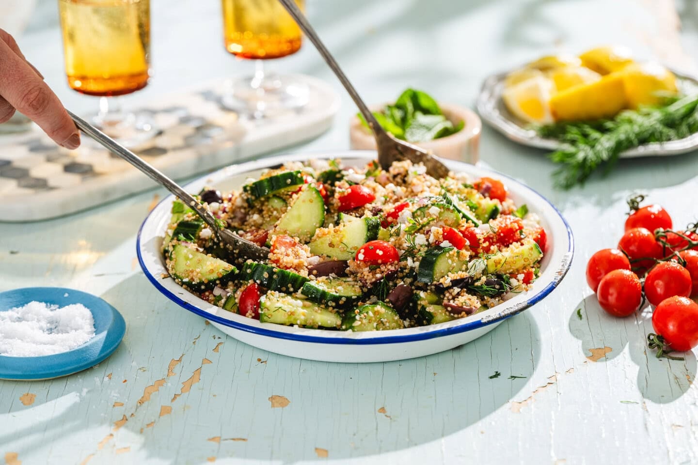 A bowl of quinoa salad with cucumbers, cherry tomatoes, and herbs is being served with tongs. Lemon wedges, cherry tomatoes, and salt are nearby on a rustic light-blue table. Two glasses of iced drinks are in the background.