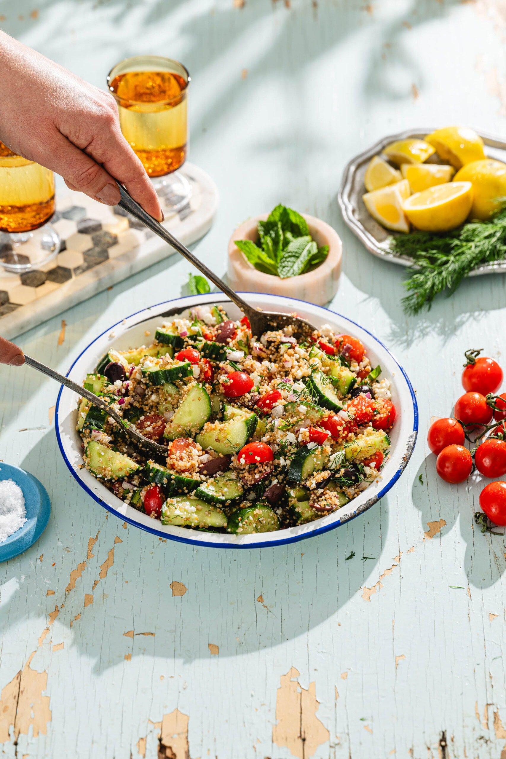 A hand tosses a fresh quinoa salad with cherry tomatoes, cucumber, and herbs in a white bowl on a rustic blue table, surrounded by lemon wedges, drinks, tomatoes, and greens.