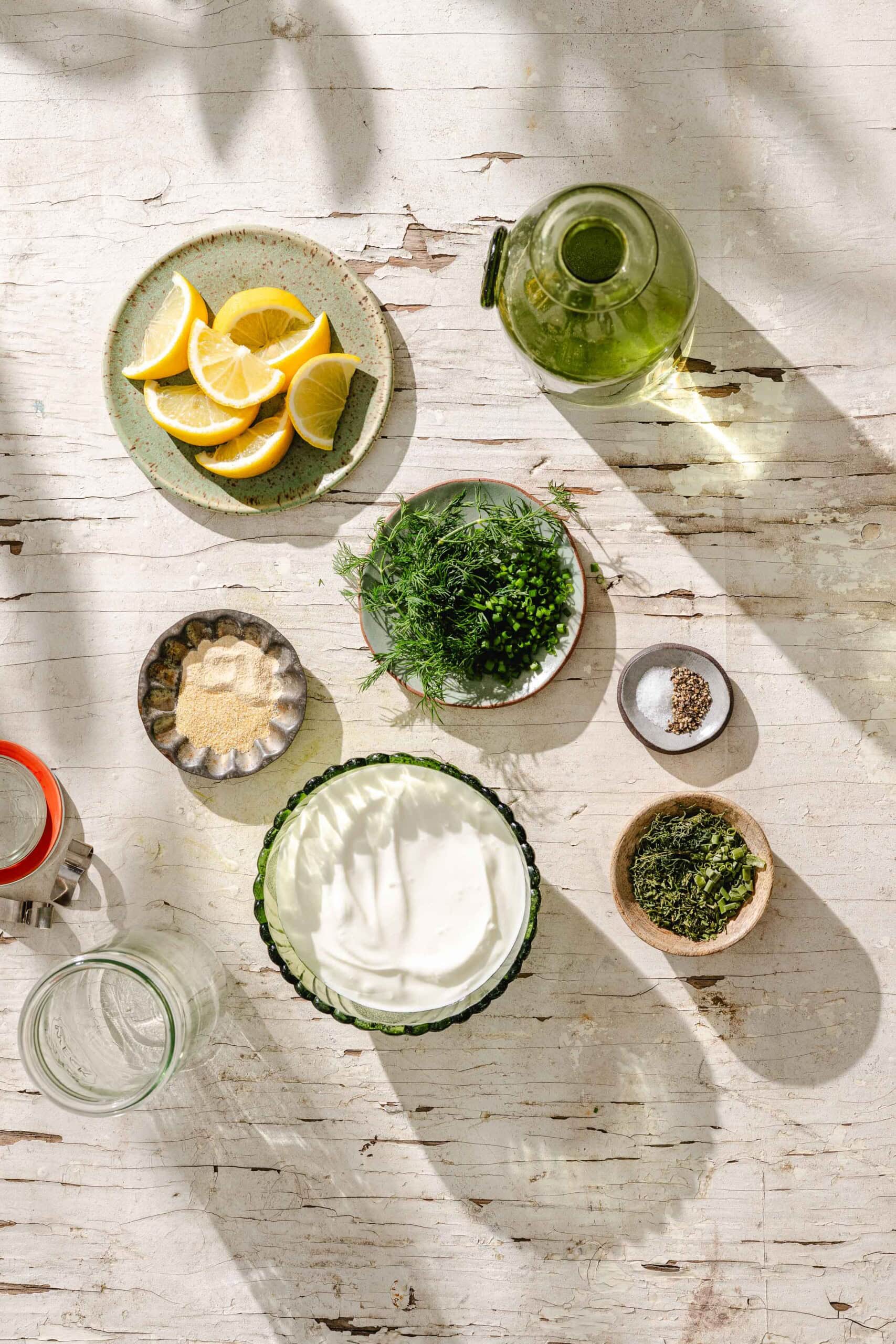 Overhead view of small bowls with lemon wedges, herbs, salt, pepper, garlic powder, a glass jar, a bowl of creamy white sauce, and a bottle of olive oil on a rustic white wooden surface.