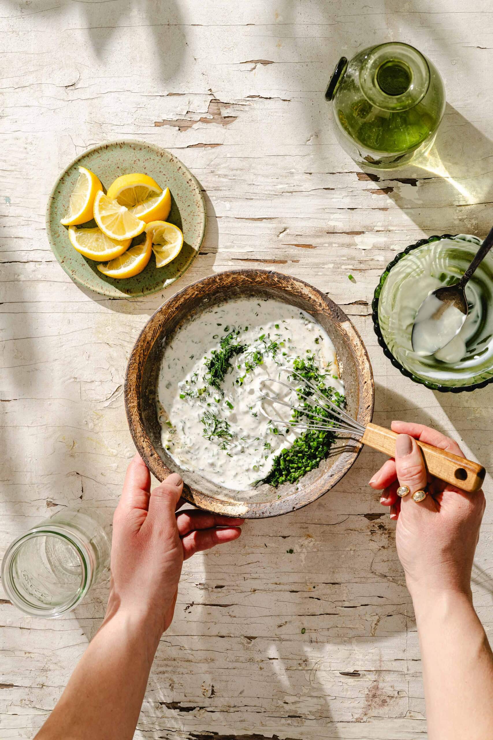 A person whisks a creamy herb sauce in a bowl on a rustic white table, surrounded by lemon wedges, a green glass bowl with a spoon, olive oil, and a glass of water.