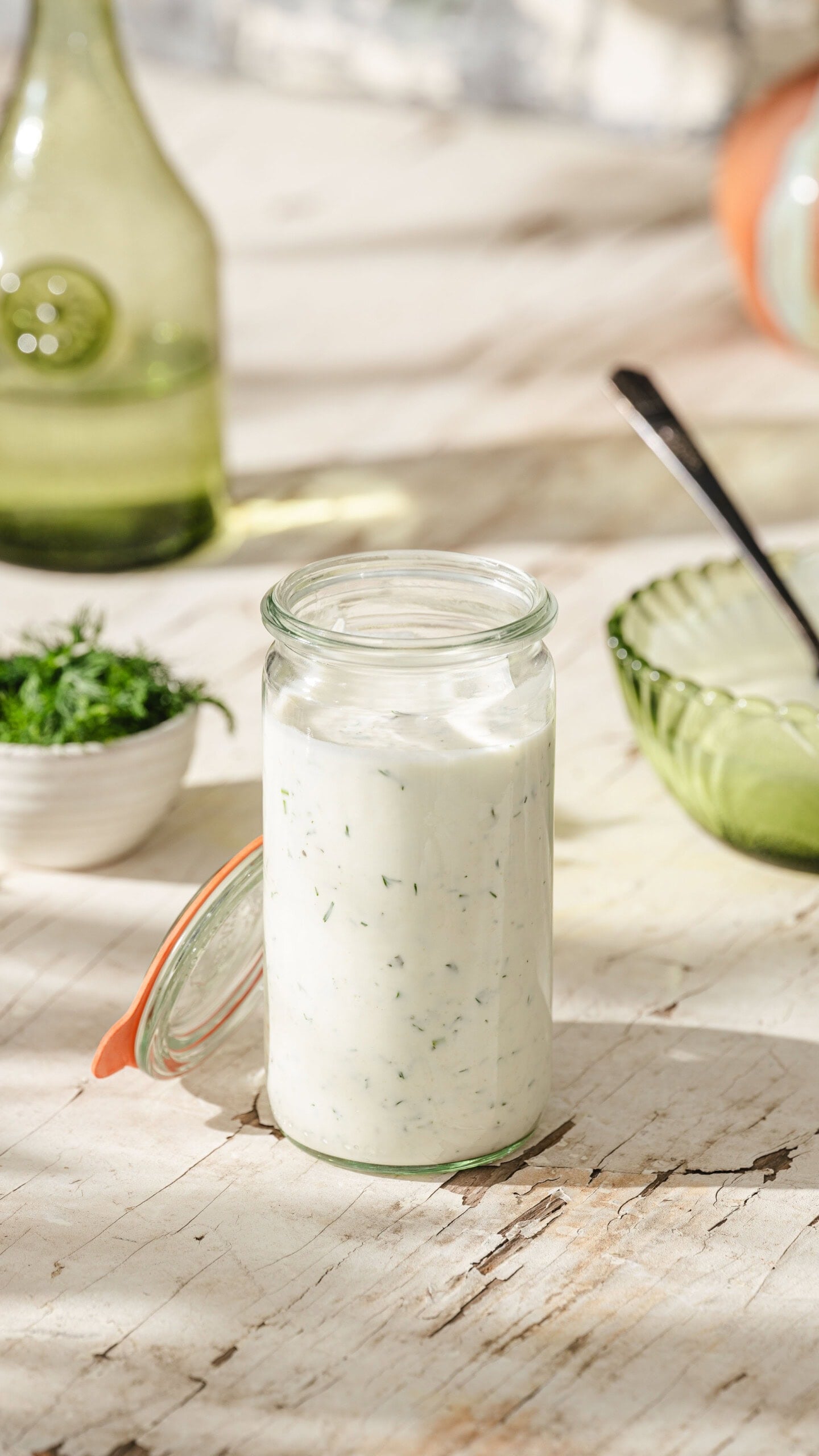A glass jar filled with creamy dressing or sauce garnished with green herbs sits on a wooden surface, surrounded by a small bowl of chopped herbs and green kitchenware in bright natural light.