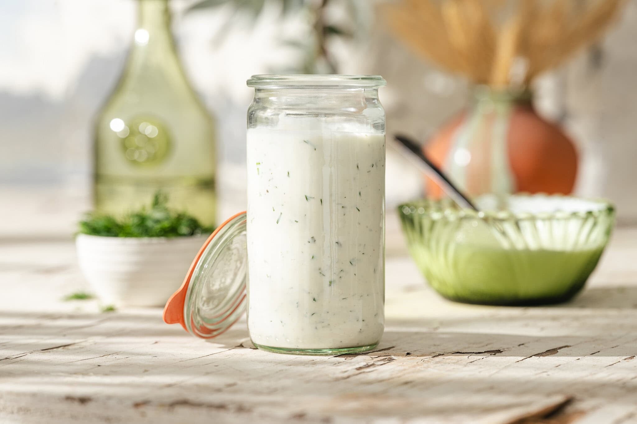 A glass jar filled with creamy white dressing with herbs, sitting on a rustic wooden table. The jars lid is open beside it. In the background are a bowl, a bottle, and blurred kitchen items.
