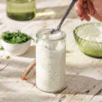 A hand holds a spoon dipping into a glass jar filled with creamy, herb-flecked ranch dressing. Fresh dill and a bowl of dressing are in the background on a light, rustic surface.