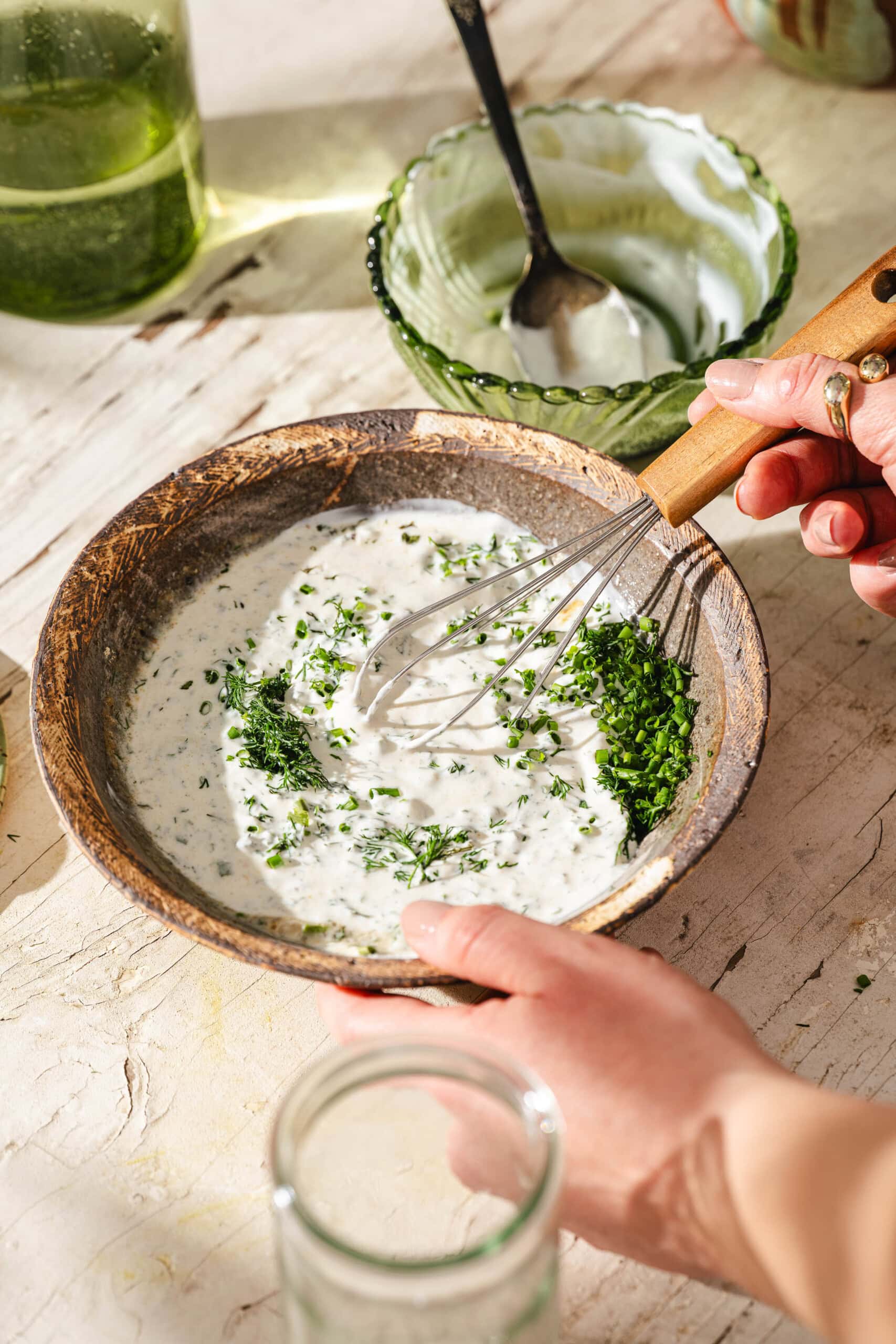 A hand holds a whisk mixing chopped herbs into a creamy sauce in a rustic bowl, with an empty green glass bowl, a spoon, and a glass jar nearby on a sunlit countertop.