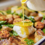 A close-up of roasted potatoes, leafy greens, and a piece of white fish topped with lemon slices, as golden sauce is poured over the fish from a small white pitcher.