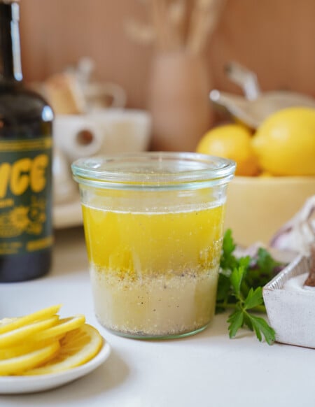 A glass jar filled with yellow salad dressing, showing separation of oil and vinegar layers, sits on a kitchen counter with sliced lemons, parsley, a bowl of lemons, and a bottle in the background.