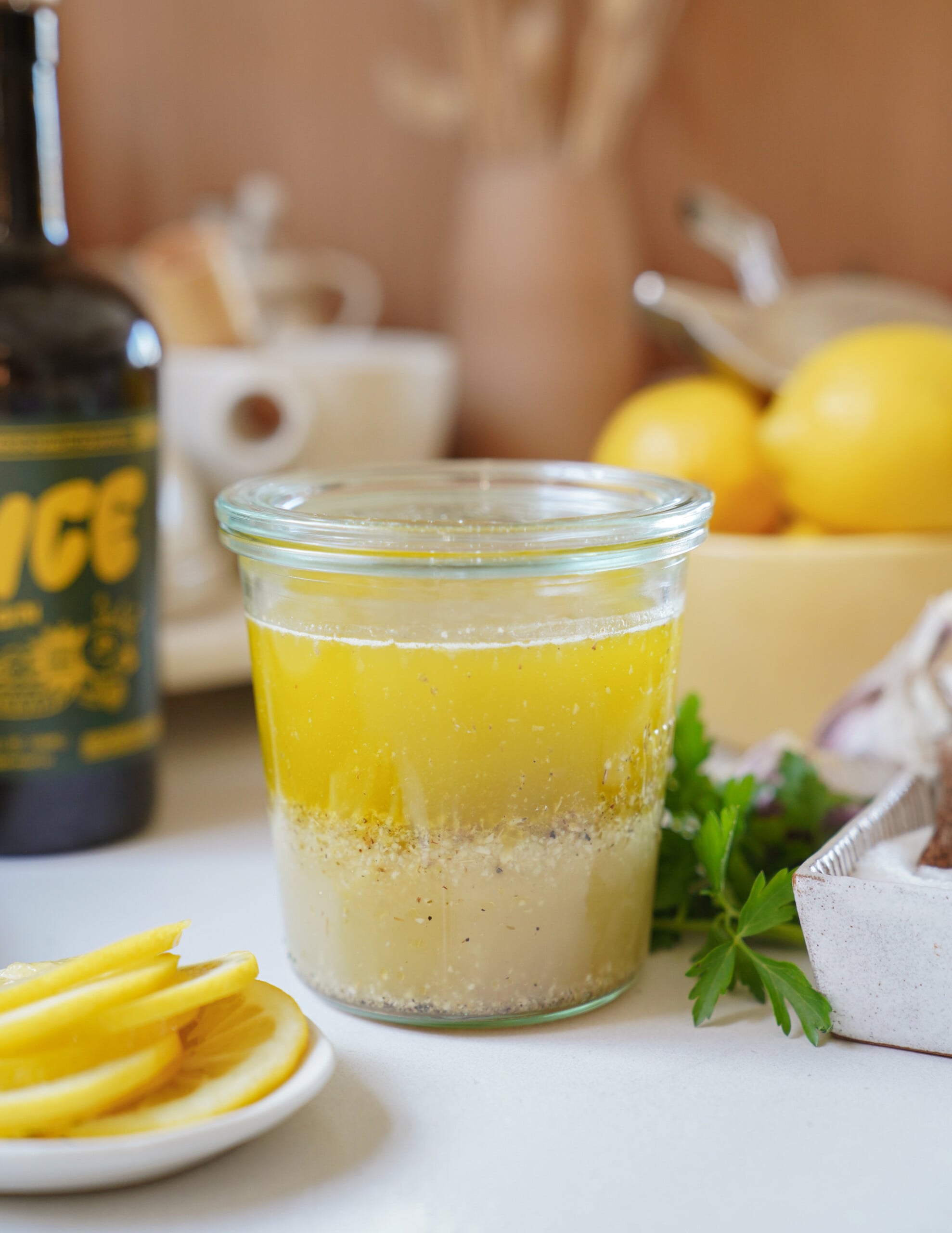 A glass jar filled with yellow salad dressing, showing separation of oil and vinegar layers, sits on a kitchen counter with sliced lemons, parsley, a bowl of lemons, and a bottle in the background.