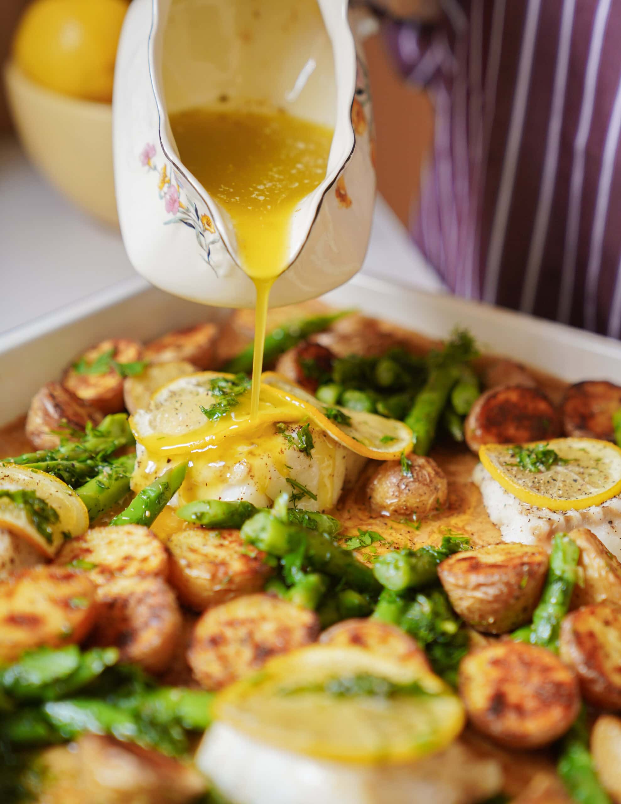 A close-up of a hand pouring yellow sauce from a floral jug onto baked fish, potatoes, asparagus, and lemon slices on a tray, creating a vibrant and appetizing meal.