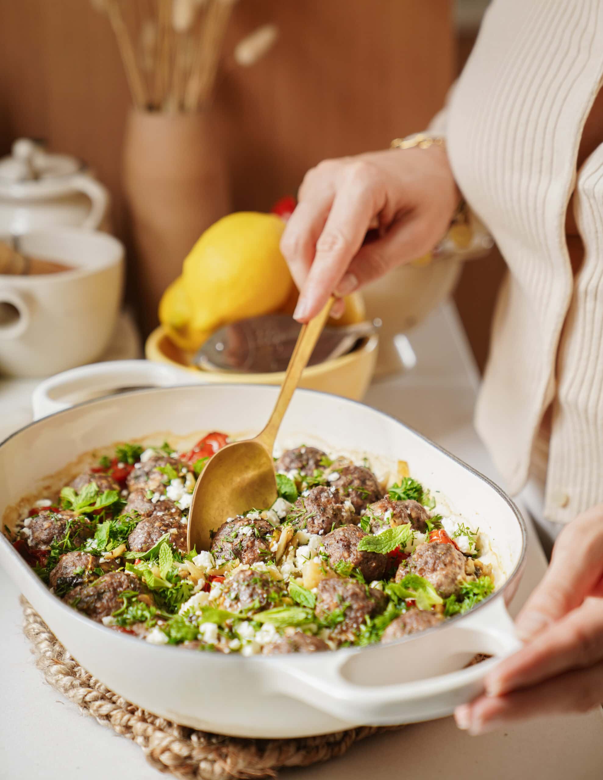 A person in a cream sweater serves a baked dish with meatballs, vegetables, and herbs from a white casserole dish using a gold spoon. A lemon and kitchenware are visible in the background.