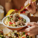 A person holds a bowl of pasta with meatballs, tomatoes, and herbs while scooping some with a gold-colored spoon. The dish appears fresh and colorful, suggesting a homemade meal.