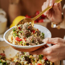 A person holds a bowl of pasta with meatballs, tomatoes, and herbs while scooping some with a gold-colored spoon. The dish appears fresh and colorful, suggesting a homemade meal.