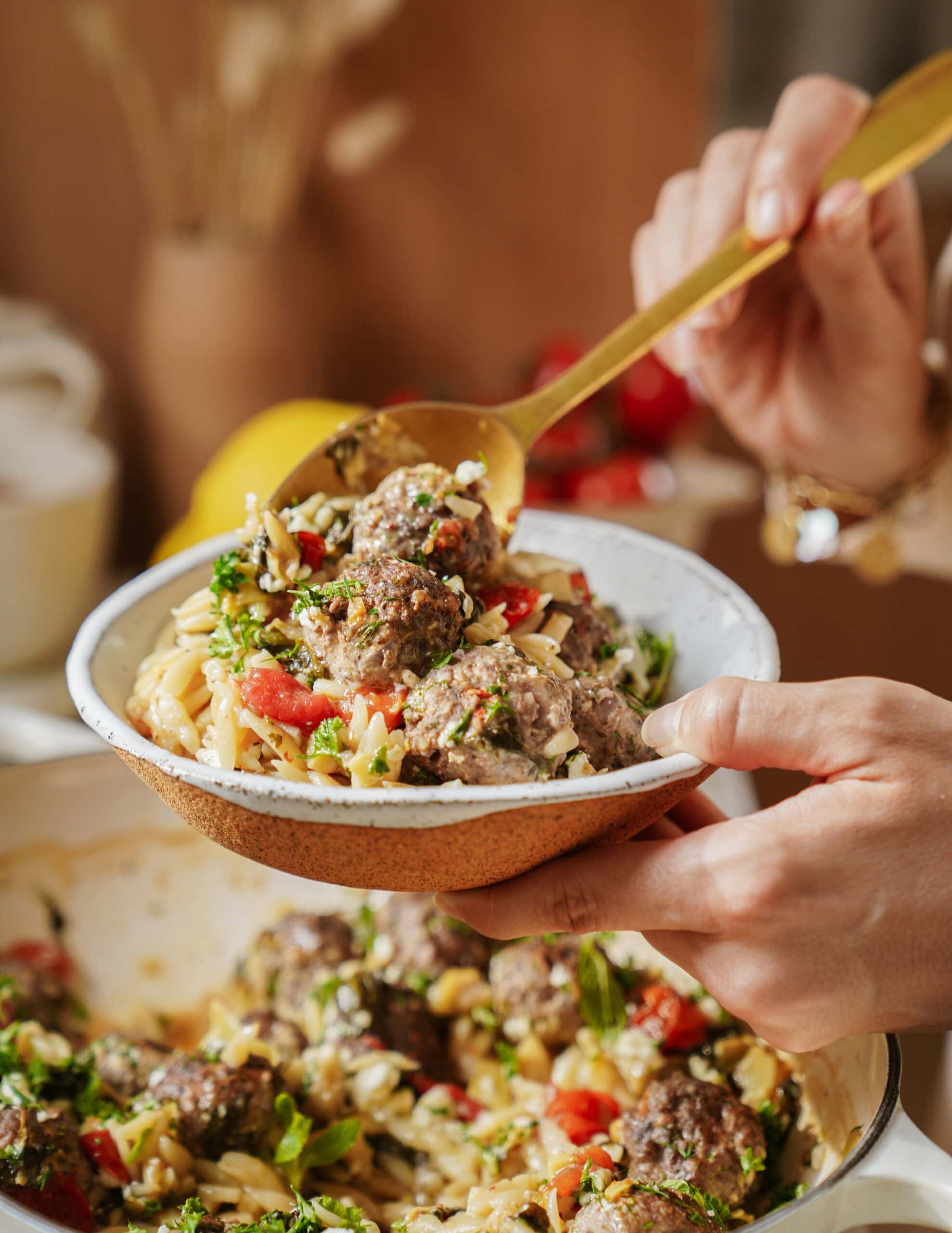 A person serves pasta with meatballs, tomatoes, and greens into a bowl using a gold spoon, with more pasta and meatballs visible in a dish below.