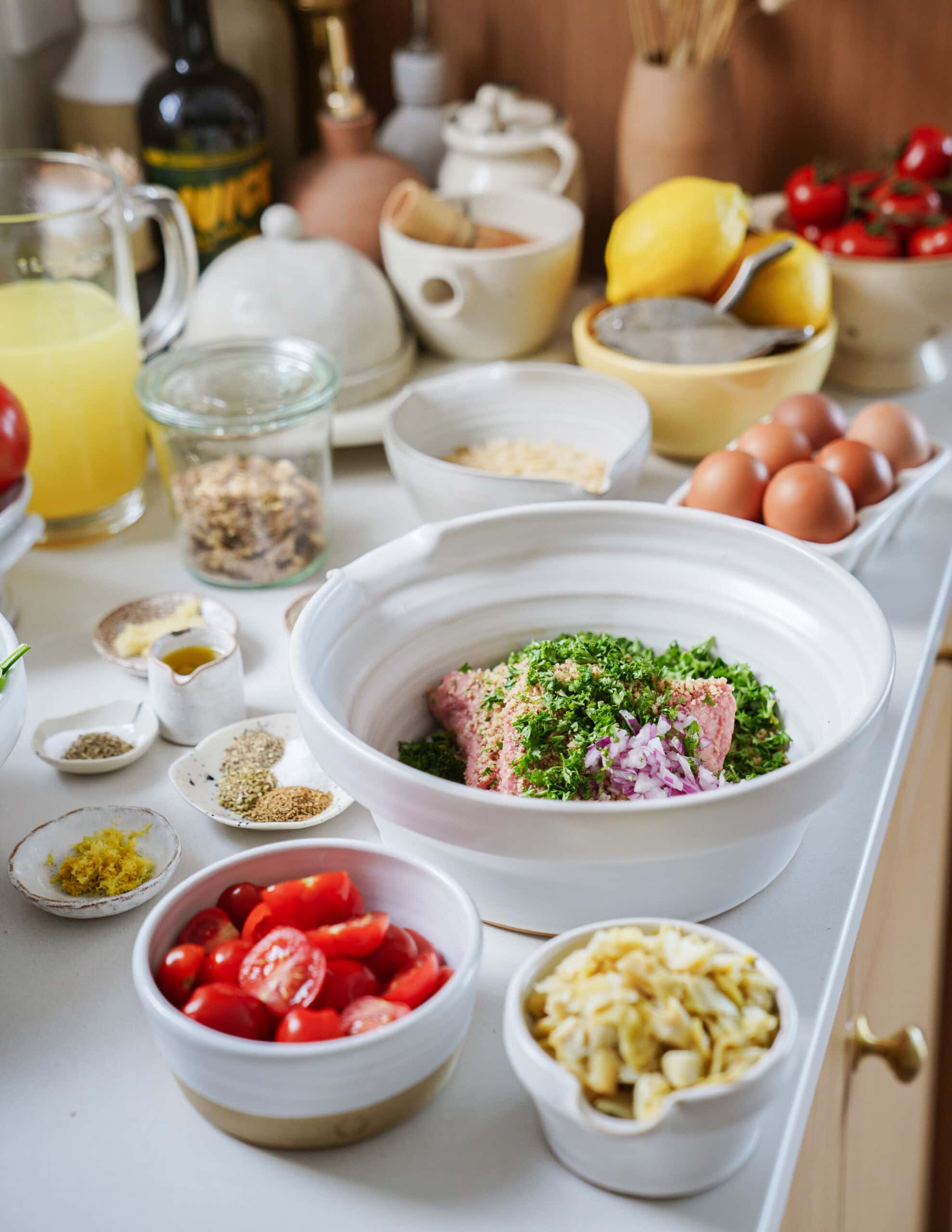 A kitchen counter with bowls of chopped tomatoes, artichokes, herbs, ground meat, onions, eggs, lemons, spices, and other cooking ingredients arranged for meal preparation.