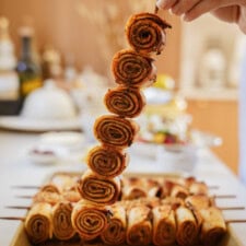 A hand holds a skewer with rolled, spiral-shaped pieces of cooked flatbread, seasoned with spices. More skewers with similar rolls are arranged on a tray in the background.