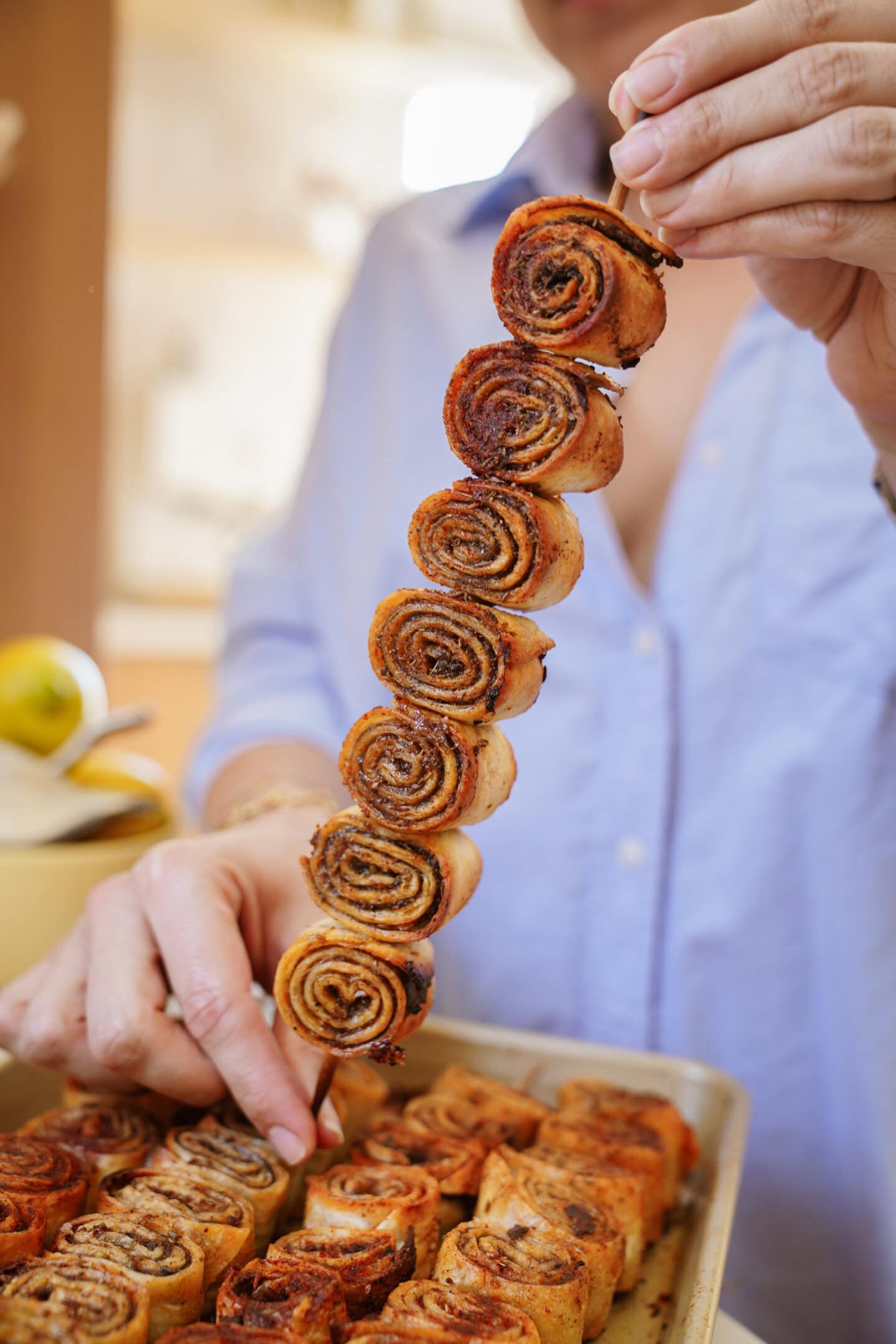 A person holds up a skewer with several small cinnamon rolls stacked on it over a tray filled with more cinnamon rolls. The rolls are golden brown and spiraled.
