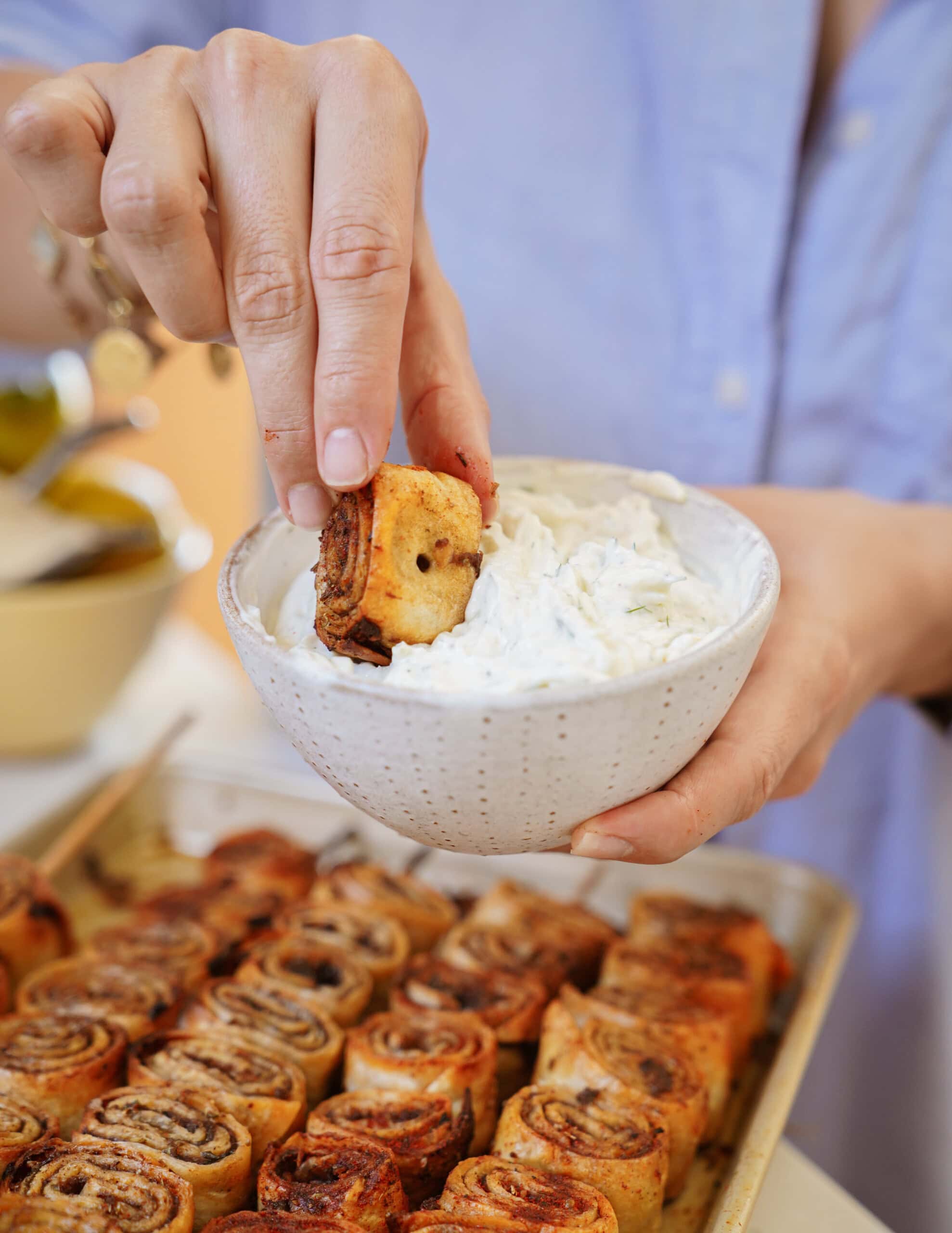 A person dips a rolled pastry into a bowl of creamy white dip, with a tray of similar pastries in the foreground. The individual is wearing a light blue shirt.
