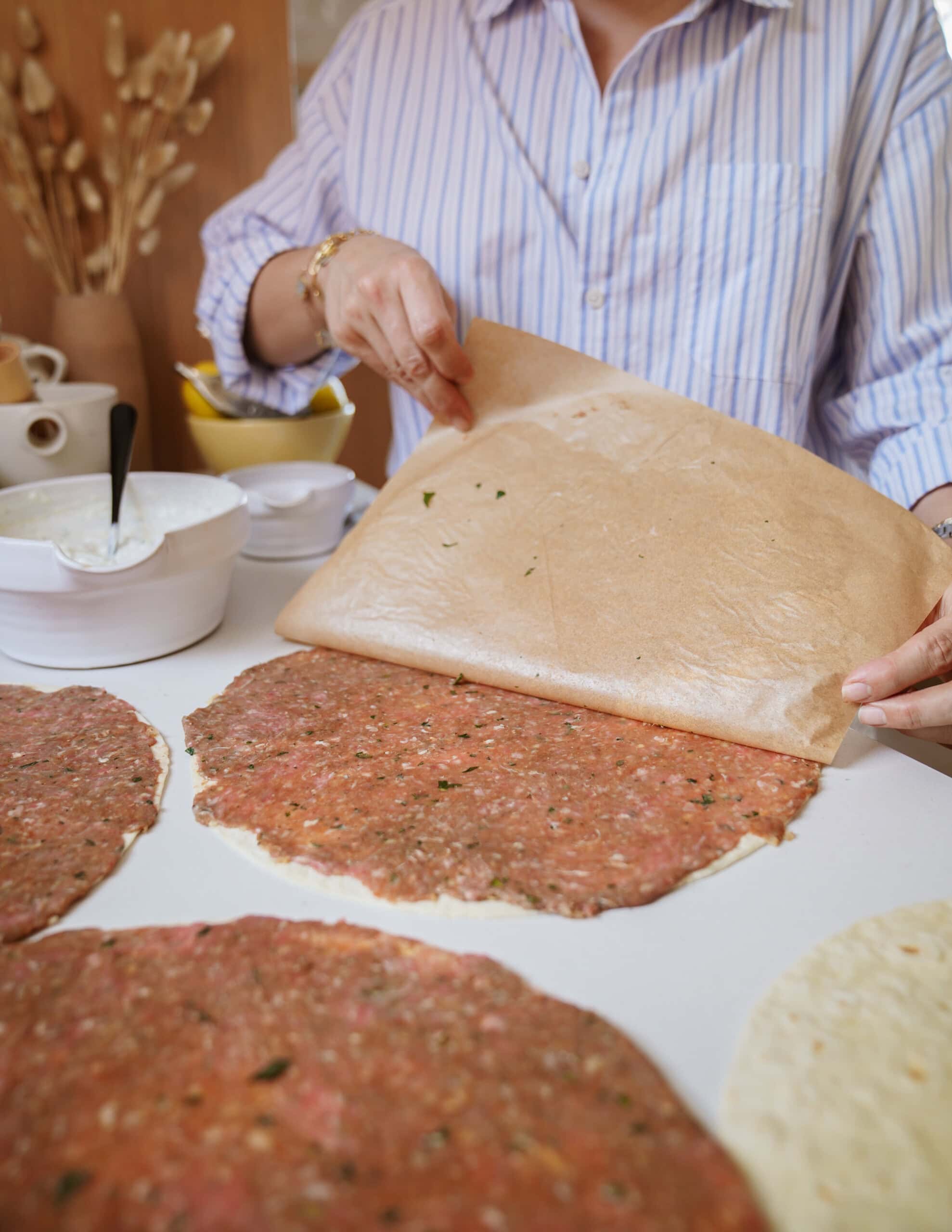 A person in a striped shirt lifts parchment paper off a round, thin layer of seasoned ground meat on a countertop. Nearby are bowls of ingredients and tools for food preparation.