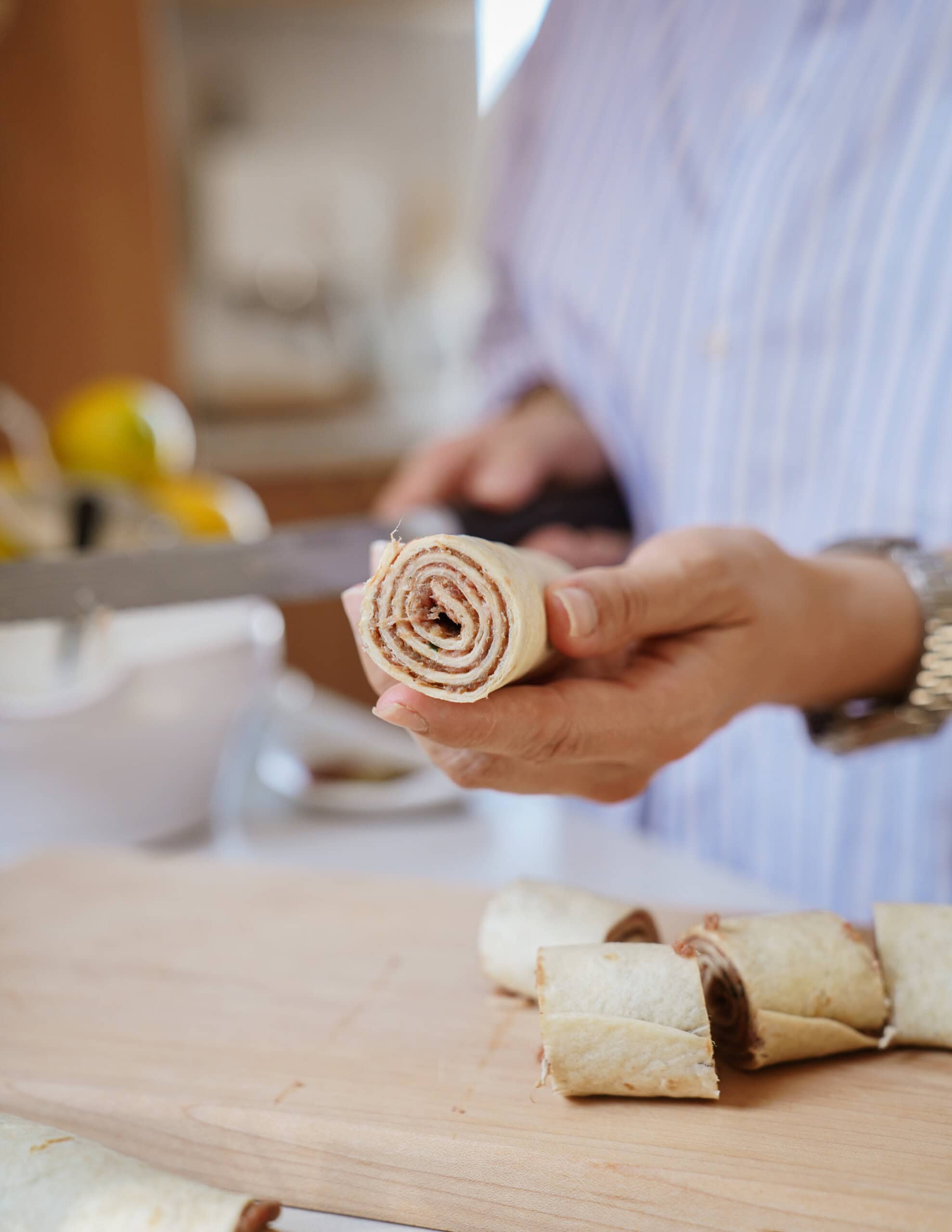 A person wearing a striped shirt holds a sliced wrap or roll, showing its spiral filling. Several more slices sit on a wooden cutting board in the foreground.