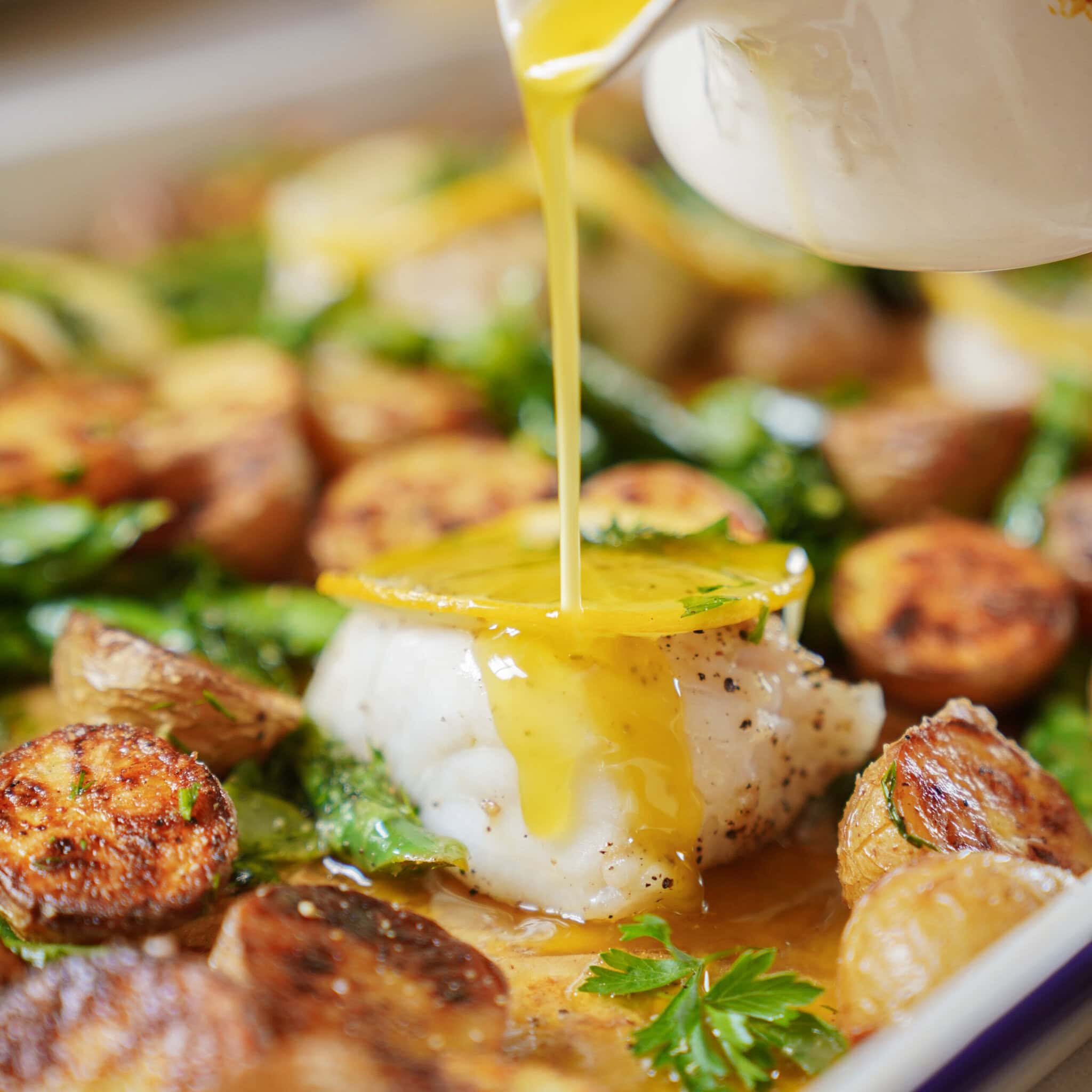 A close-up of roasted potatoes and leafy greens surrounding a piece of white fish topped with a lemon slice, as golden sauce is being poured over it. Parsley garnishes the dish.