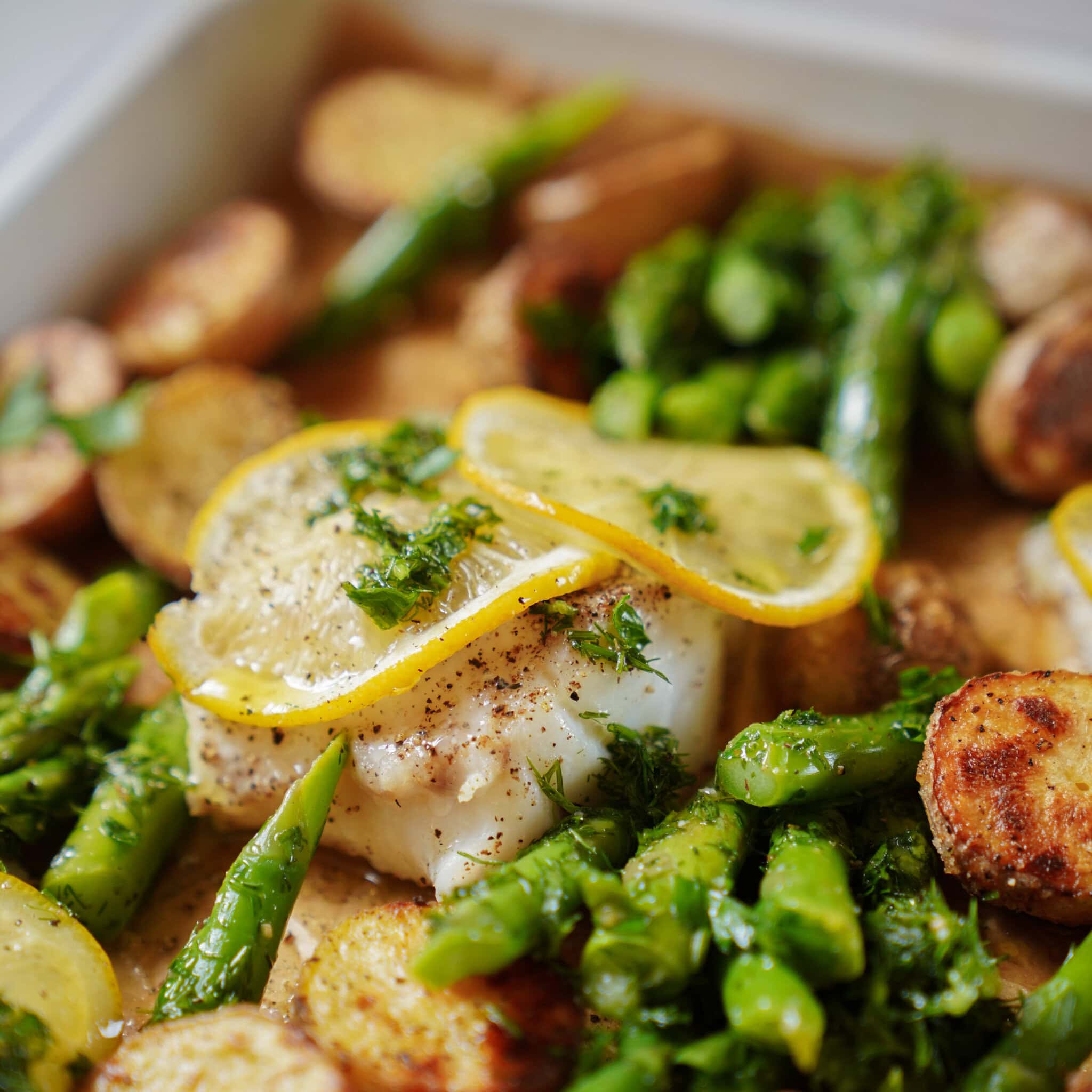 Close-up of a baked fish fillet topped with lemon slices, surrounded by roasted potato slices, asparagus, and fresh herbs on a baking tray. The dish looks vibrant and freshly cooked.