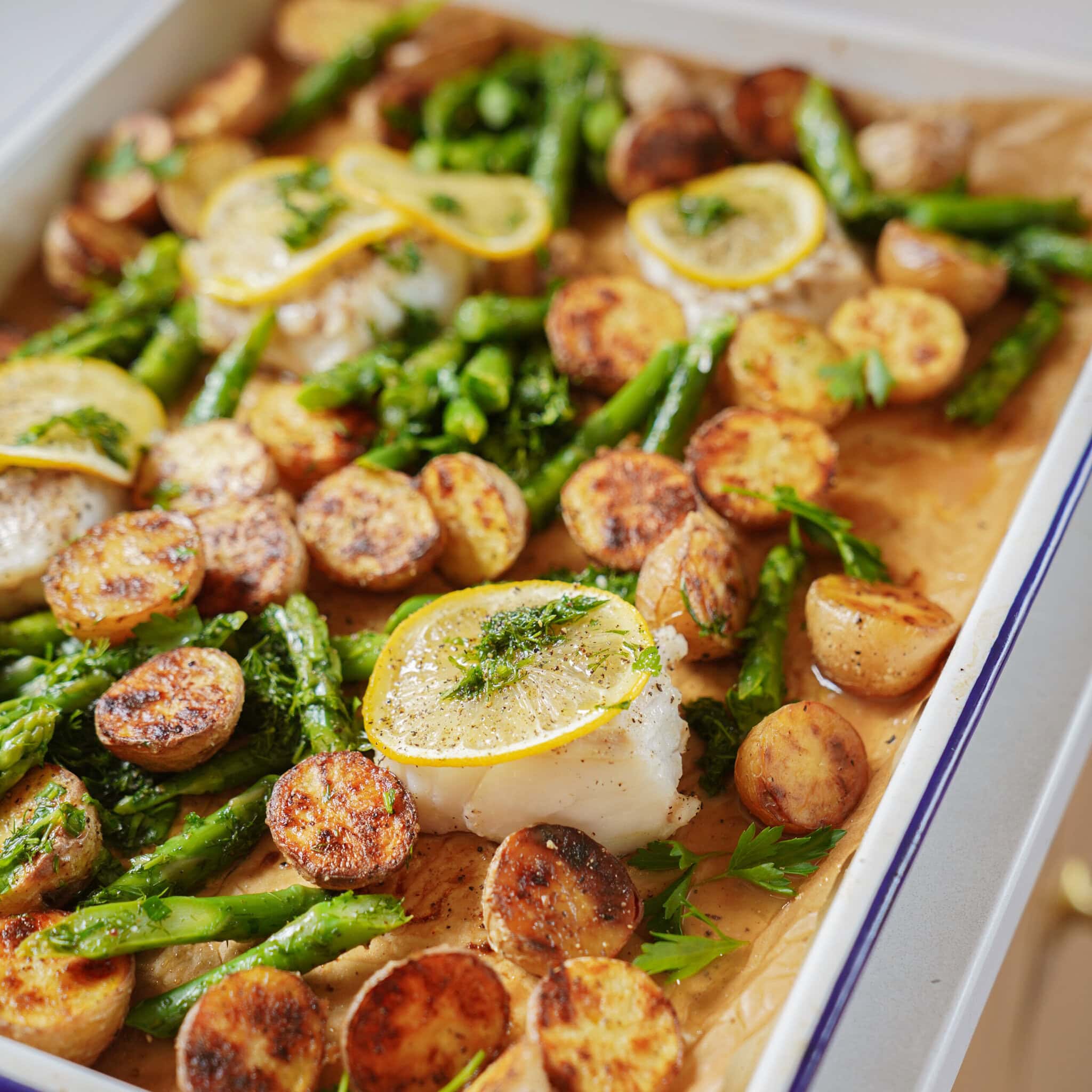 Sheet pan with roasted asparagus, sliced baby potatoes, and white fish fillets topped with lemon slices and fresh herbs, all on parchment paper. The food looks golden and freshly baked.