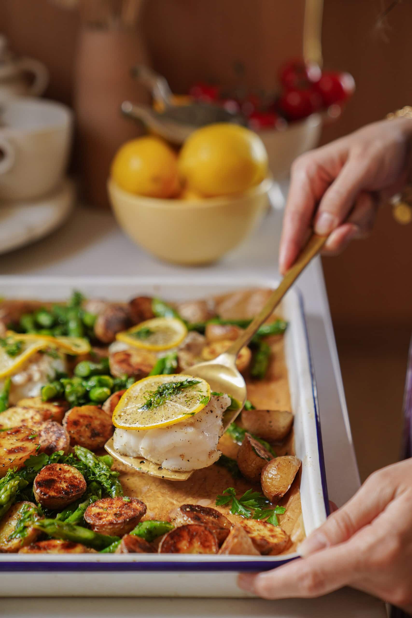 A person serves baked white fish topped with lemon and herbs from a tray with roasted potatoes, asparagus, and greens. In the background, a bowl of lemons and a few cherries are visible.