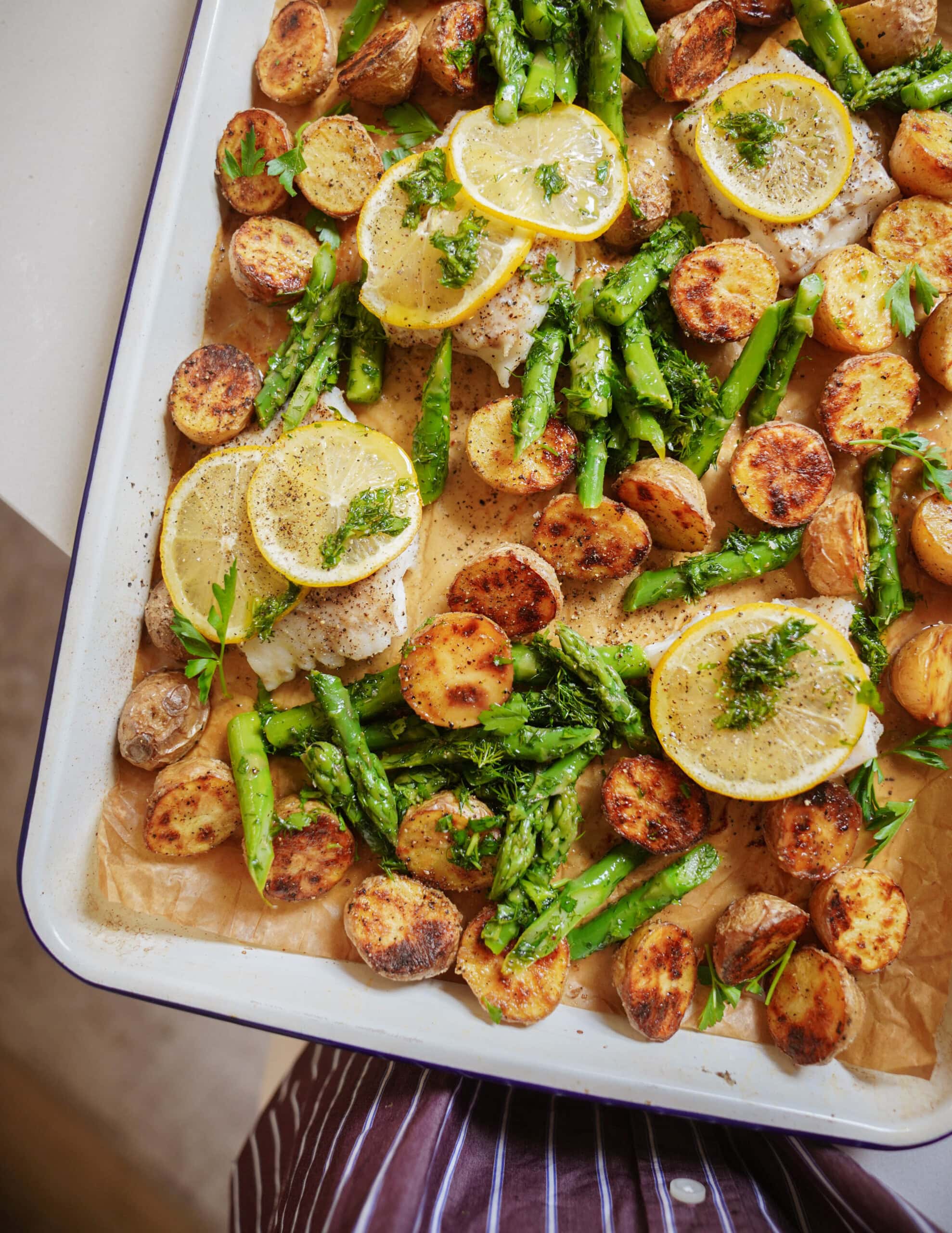 A baking tray filled with roasted potatoes, asparagus, and fish fillets topped with lemon slices and fresh herbs, all arranged on parchment paper.