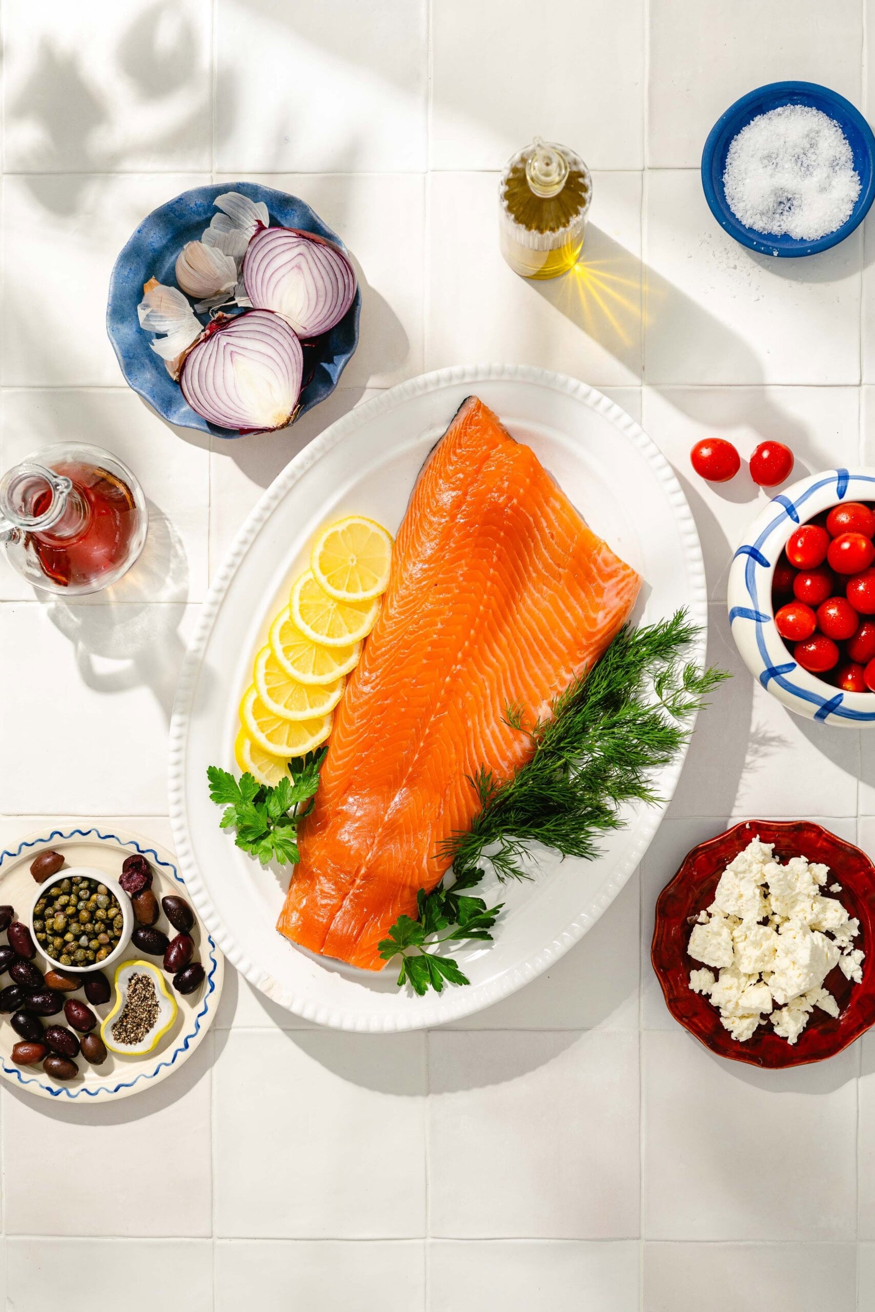 A large fillet of raw salmon on a white platter with lemon slices and herbs, surrounded by bowls of feta cheese, cherry tomatoes, olives, capers, figs, red onion, garlic, olive oil, and salt on a tiled surface.
