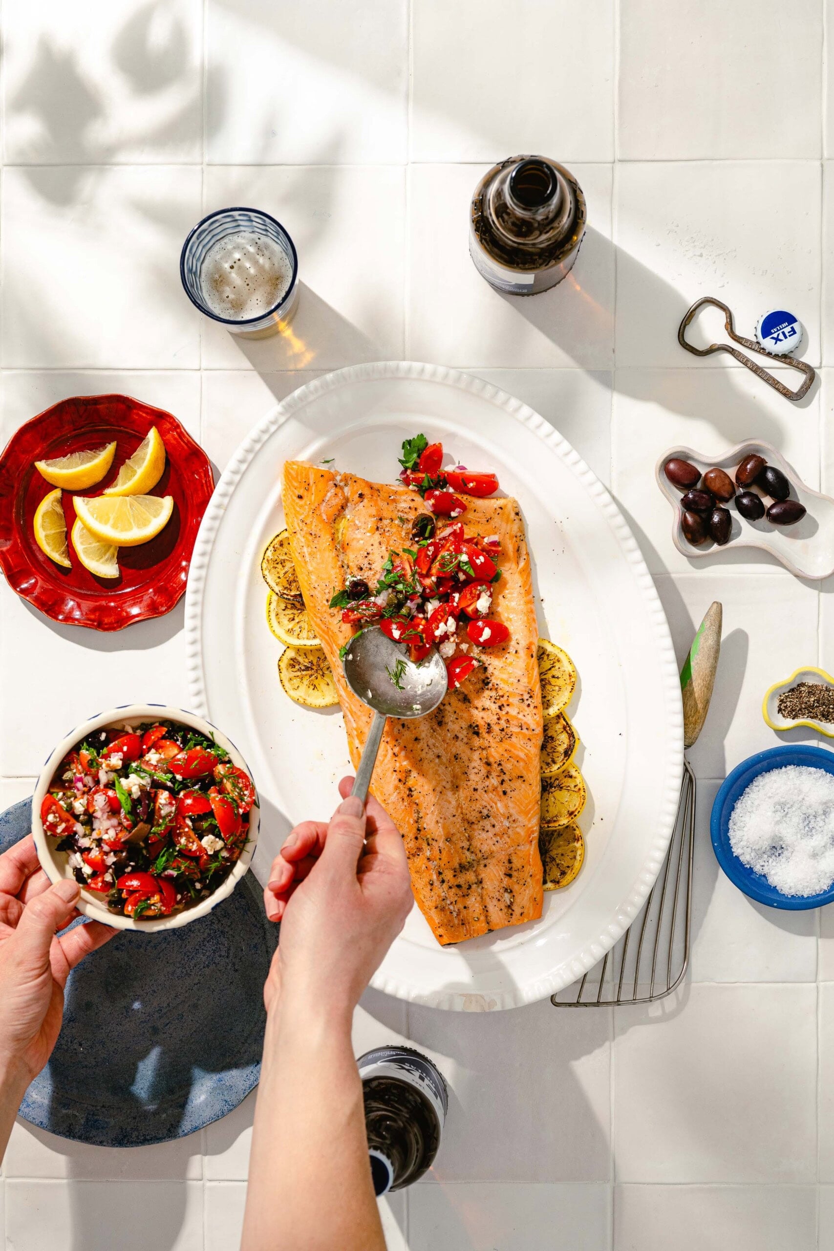 A hand spoons a fresh tomato and herb salsa over a roasted salmon fillet on a platter with grilled lemon slices, surrounded by lemon wedges, olives, a drink, a bottle, and salt on a white tiled table.