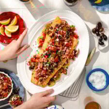 A person serves a platter of baked salmon topped with a colorful tomato, olive, and herb salsa, surrounded by lemon slices. The table is set with lemons, olives, salt, and side dishes.