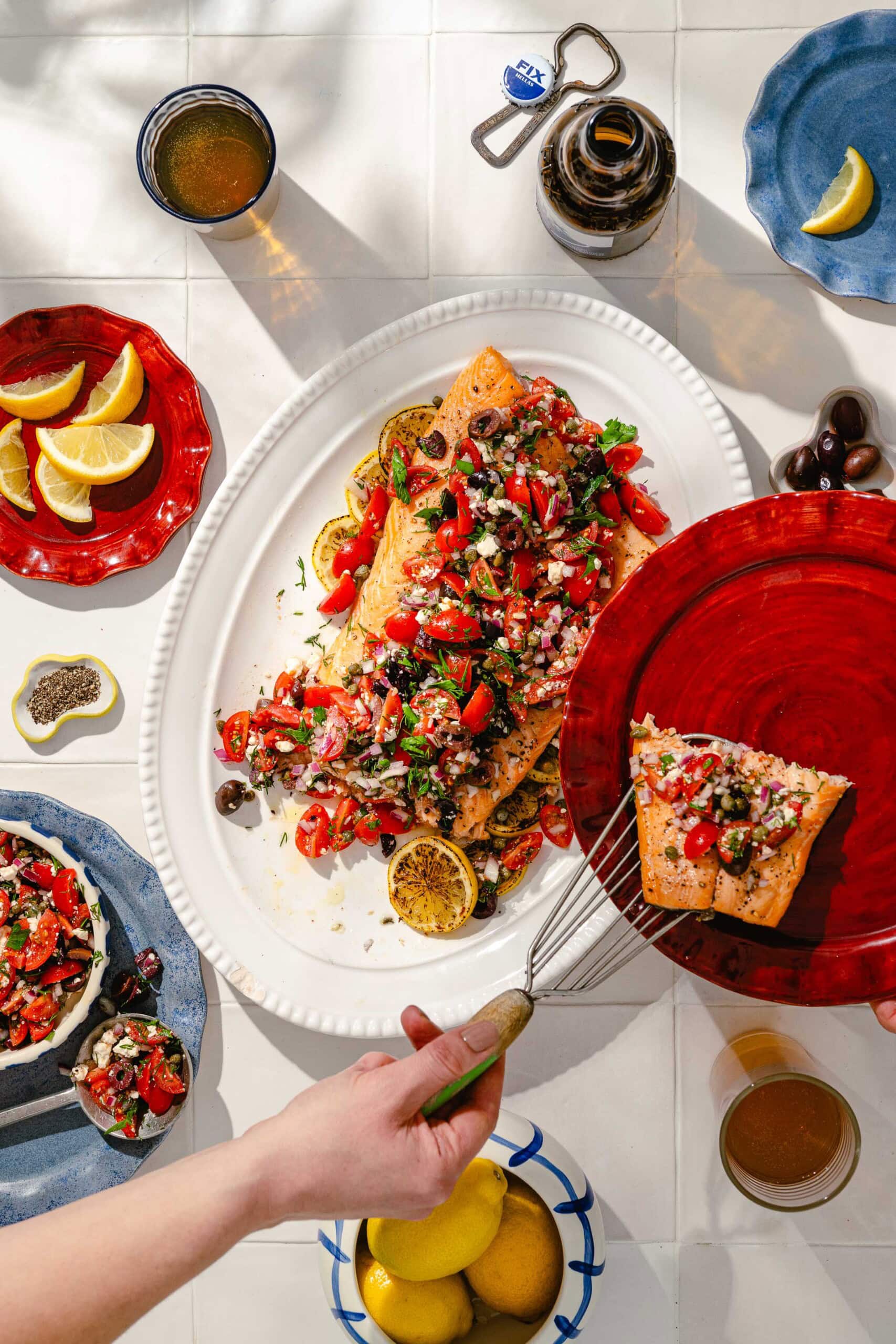 A hand serves a slice of baked salmon topped with a colorful tomato and olive salsa from a white platter. Surrounding dishes hold lemon wedges, olives, drinks, and more salsa, all arranged on a white tiled surface.
