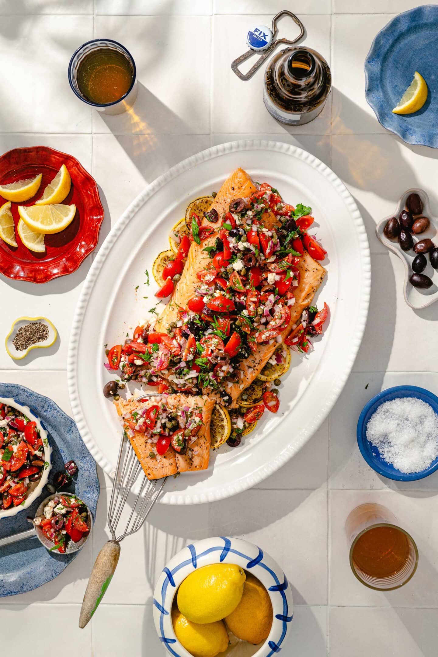 A platter of baked salmon topped with a colorful tomato, onion, and olive salsa sits on a white table surrounded by lemon wedges, drinks, olives, salt, pepper, and a plate of extra topping.