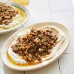 A plate topped with creamy yogurt, olive oil, and a colorful mixture of finely chopped vegetables and herbs, likely a Mediterranean or Middle Eastern salad, set on a white tiled surface.