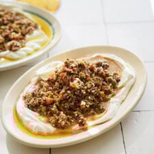 A plate topped with creamy yogurt, olive oil, and a colorful mixture of finely chopped vegetables and herbs, likely a Mediterranean or Middle Eastern salad, set on a white tiled surface.
