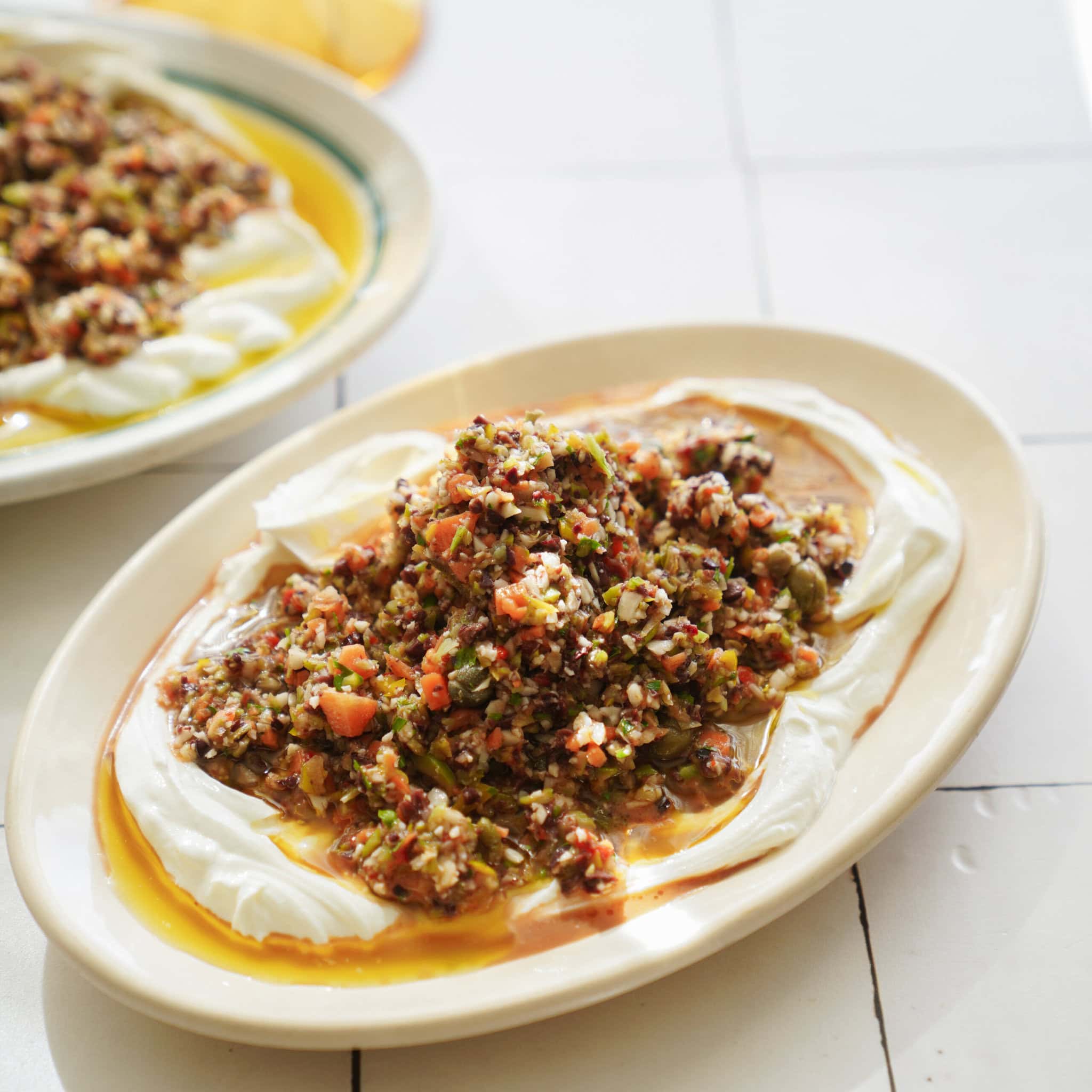 A plate topped with creamy yogurt, olive oil, and a colorful mixture of finely chopped vegetables and herbs, likely a Mediterranean or Middle Eastern salad, set on a white tiled surface.