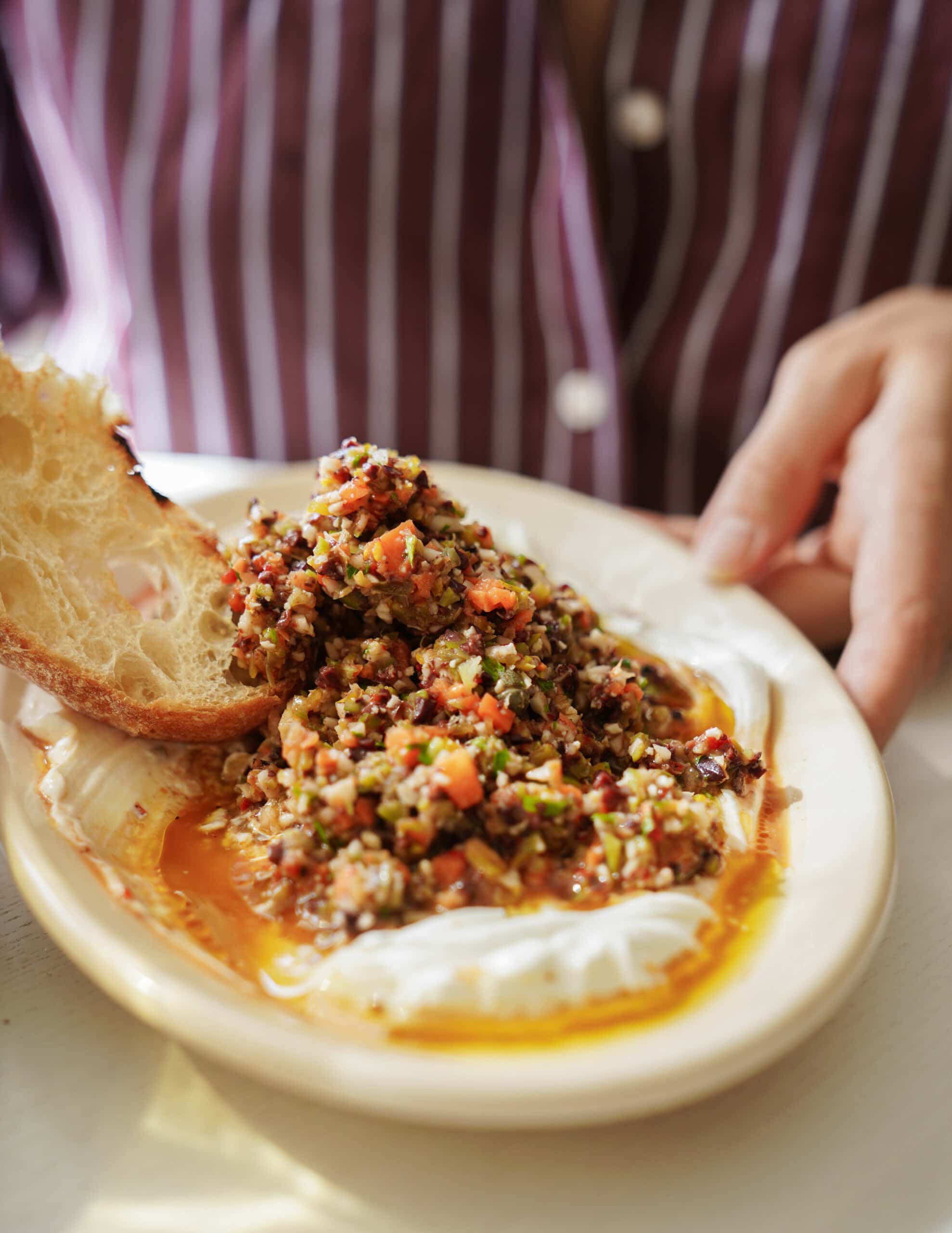 A hand holds a plate with a slice of bread, creamy spread, and a colorful chopped vegetable mix, drizzled with olive oil. The person wears a maroon and white striped shirt.