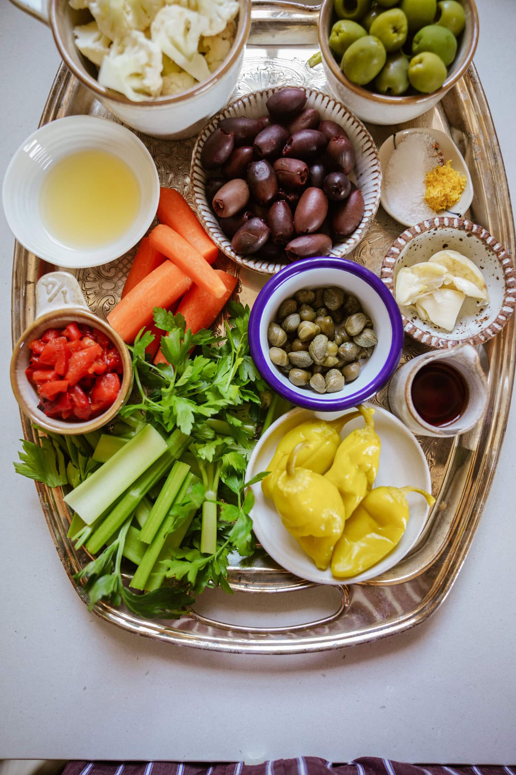 A silver tray with bowls of green and black olives, lemon juice, grated garlic, capers, pepperoncini, chopped red peppers, carrot sticks, celery, parsley, and lemon wedges arranged neatly.