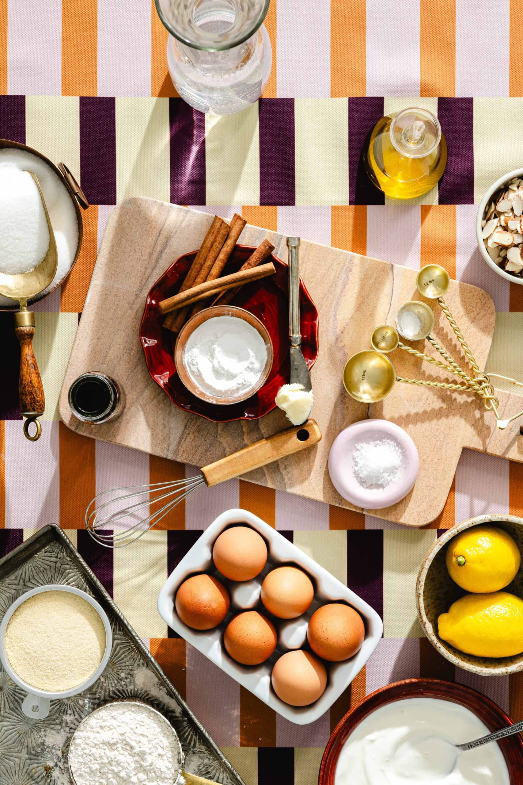 A colorful overhead view of baking ingredients, including eggs, lemons, flour, sugar, cinnamon sticks, vanilla, butter, olive oil, nuts, measuring spoons, and a whisk arranged on a striped patterned tablecloth.