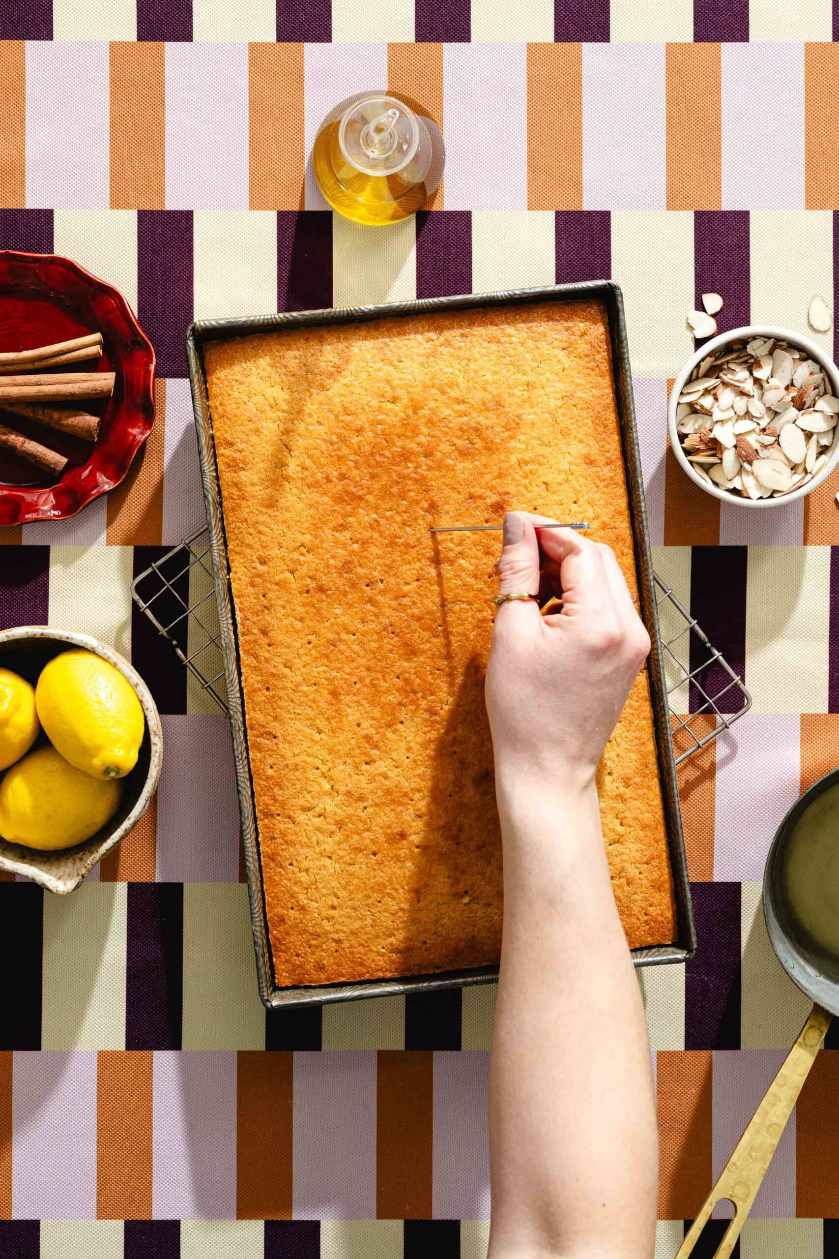 A hand slices into a rectangular golden cake in a baking pan. Around it are bowls with cinnamon sticks, almond slices, lemons, syrup in a pan, and a bottle of oil. The background is a patterned tablecloth.