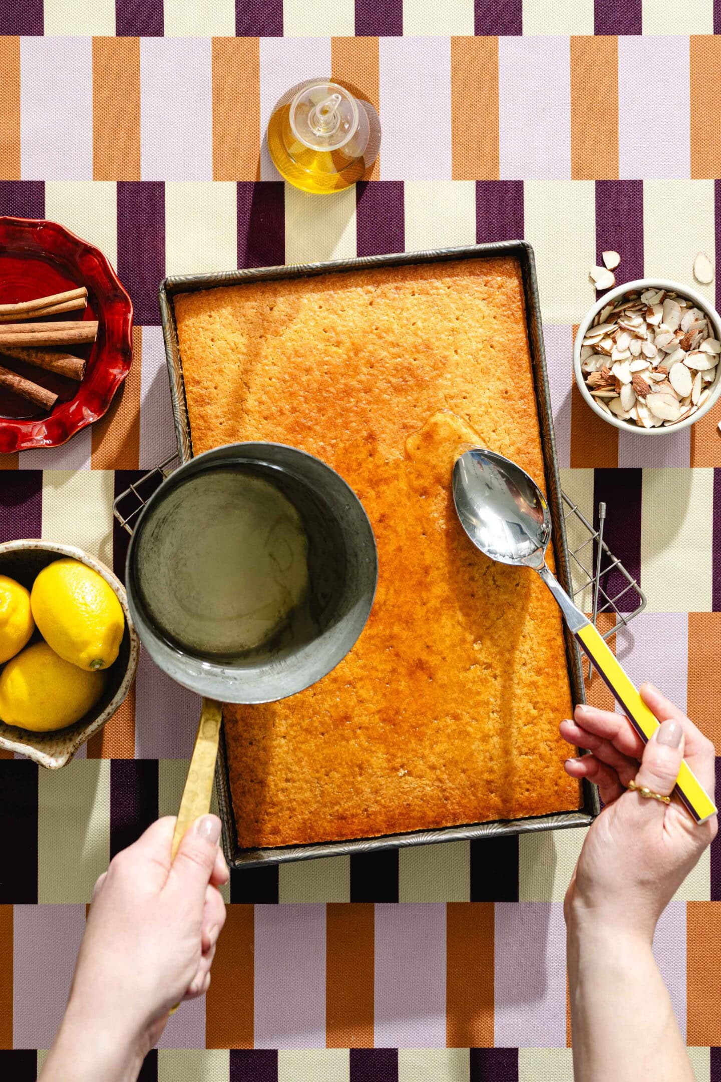 A person pours syrup from a saucepan onto a rectangular cake using a ladle. Surrounding the cake are lemons, cinnamon sticks, sliced almonds, and a small bottle of oil on a patterned tablecloth.