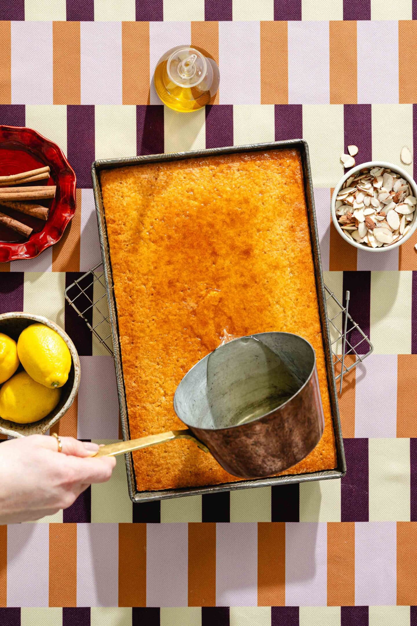 A hand pours syrup from a saucepan onto a rectangular golden cake in a pan. Nearby are lemons, cinnamon sticks, sliced almonds, and a bottle of syrup on a patterned tablecloth.