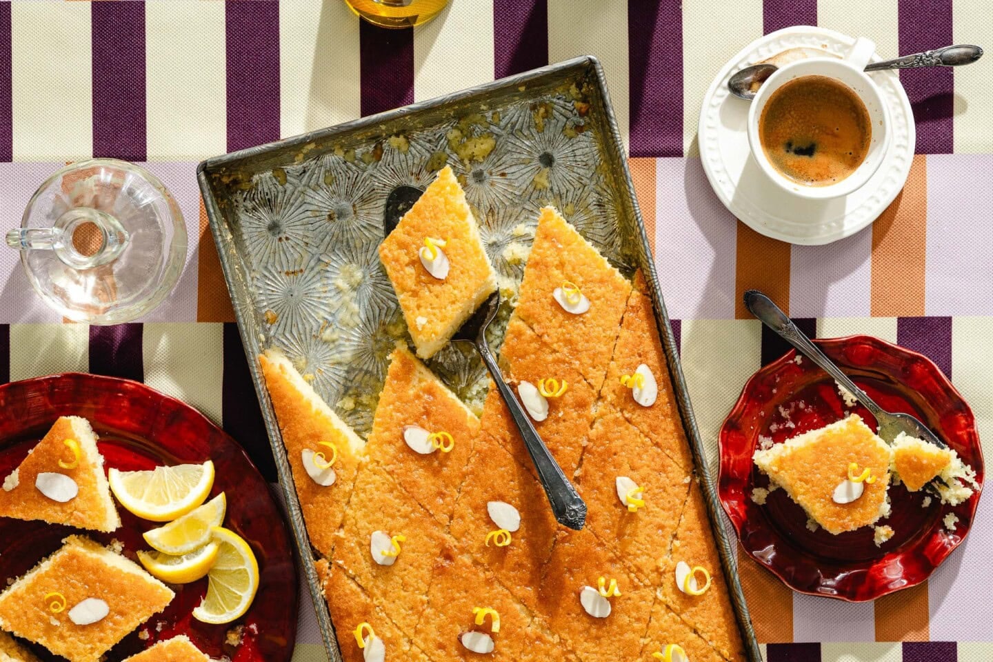 A rectangular tray of semolina cake decorated with almond slices and lemon zest sits on a striped tablecloth, with slices served on red plates, lemon wedges, a cup of coffee, and a glass of water nearby.