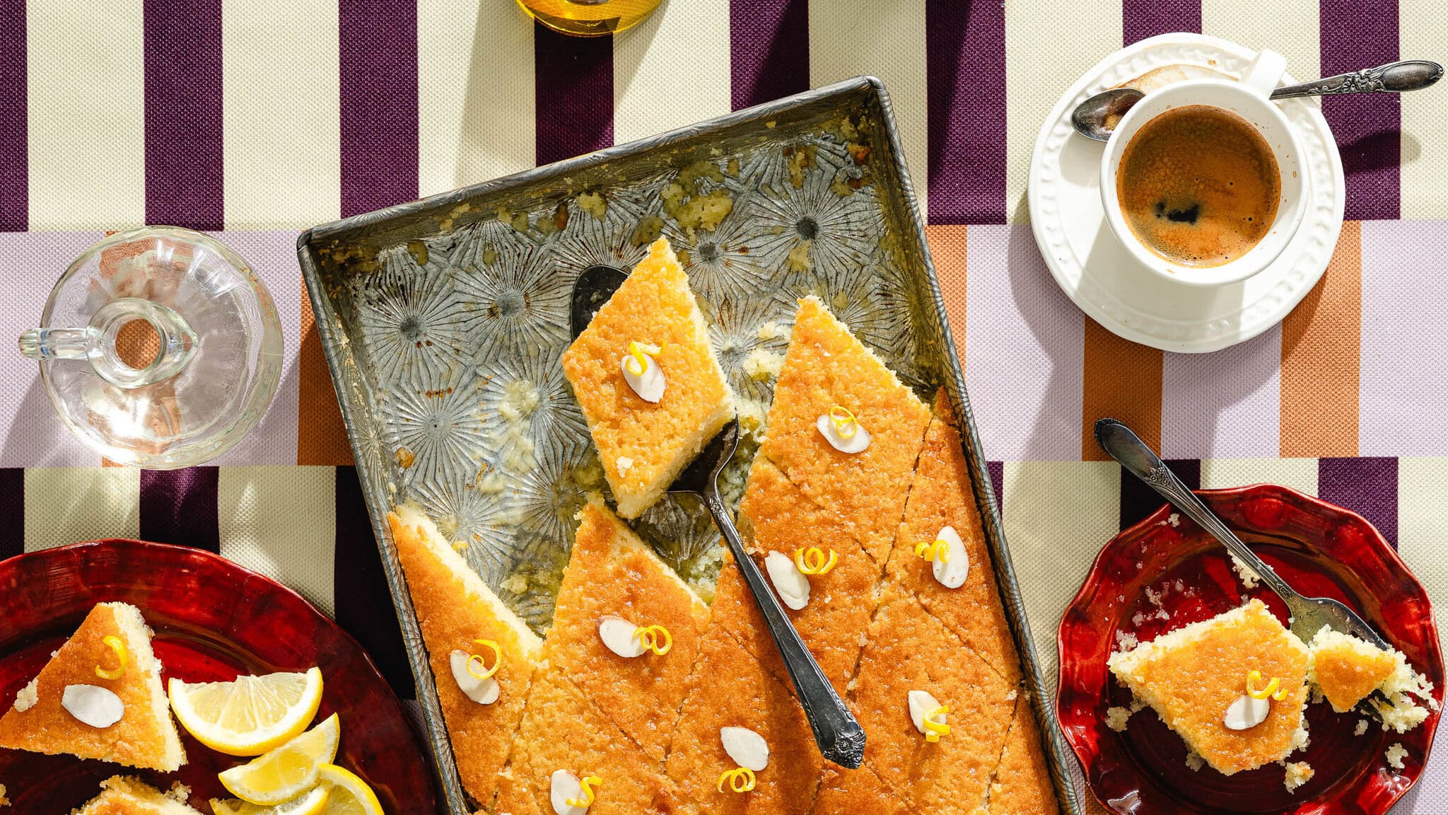 A tray of diamond-shaped lemon cake slices garnished with almond slivers and lemon zest sits on a striped tablecloth, surrounded by plates with cake slices, lemon wedges, a cup of coffee, and a glass jug of water.