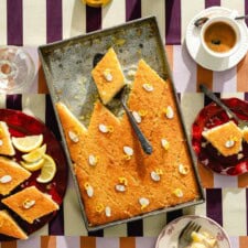 A tray of golden, diamond-shaped lemon cake slices garnished with almond slivers and lemon zest, surrounded by sliced lemons, coffee, and water on a striped tablecloth.