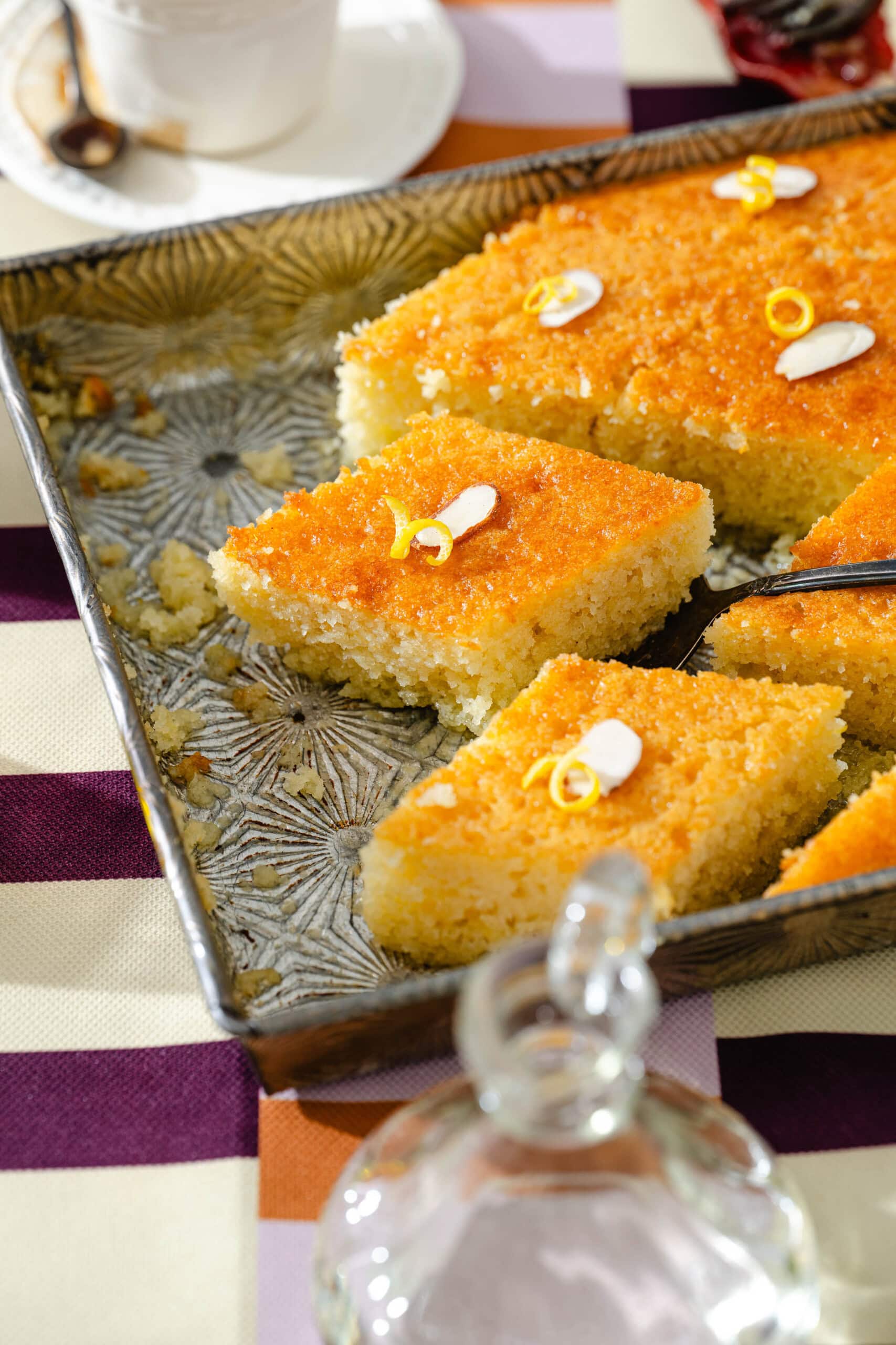 A tray of golden, syrup-soaked cake pieces, each topped with almond slivers and orange zest, sits on a patterned surface with a glass container and a teacup in the background.