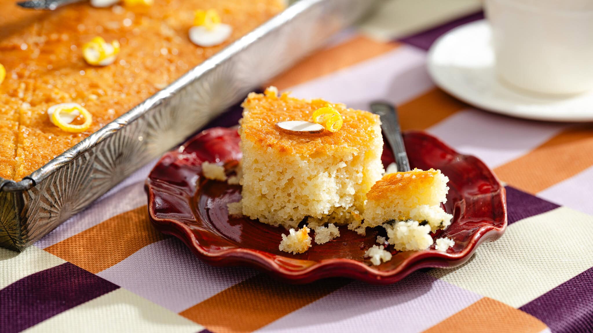 A square piece of golden, moist cake topped with nuts sits on a red plate with crumbs and a fork, next to a baking tray of more cake on a colorful checkered tablecloth.
