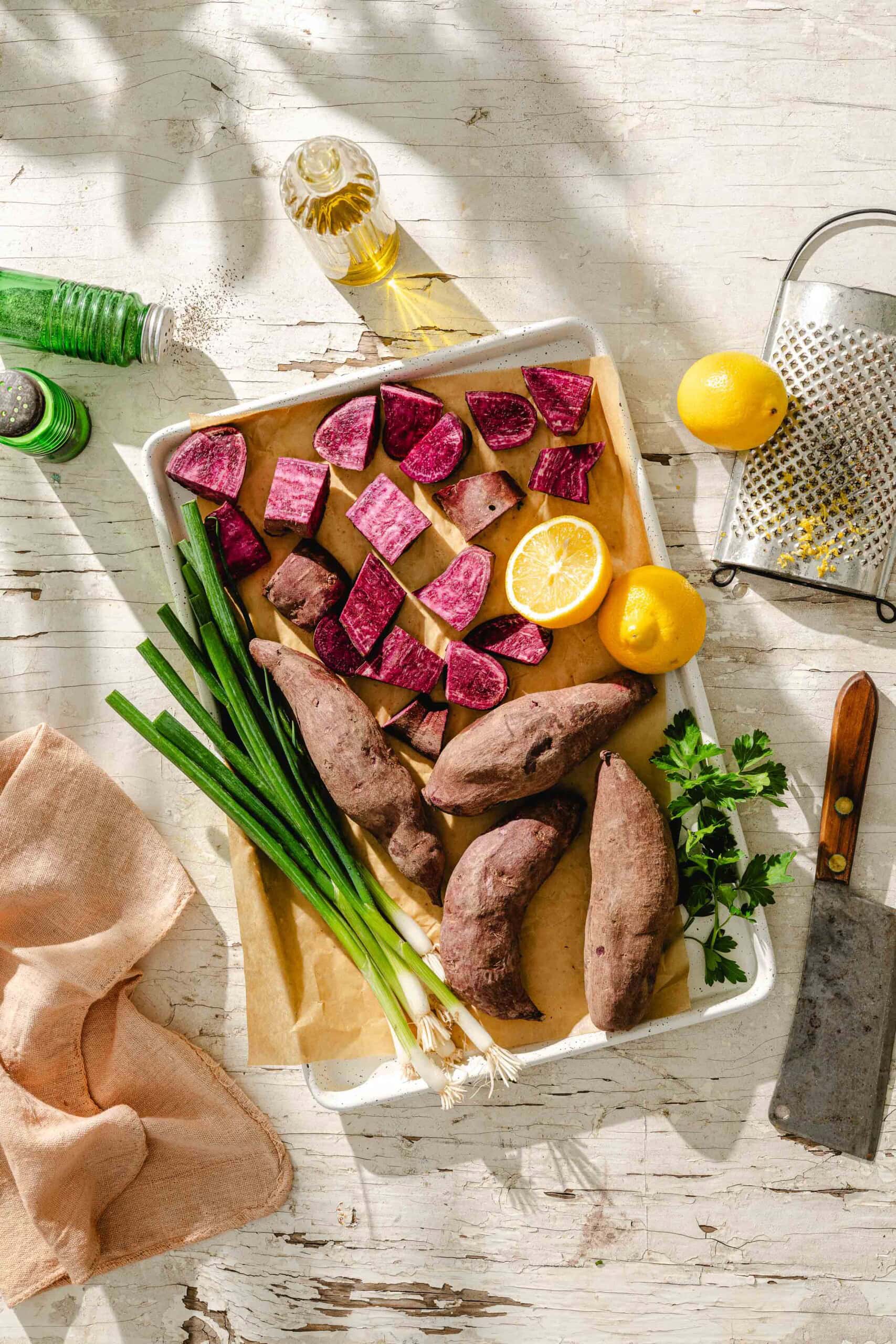A baking tray with whole and chopped purple sweet potatoes, a halved lemon, green onions, and parsley on parchment paper, surrounded by a grater, knife, oil bottle, green spice shaker, and a peach cloth on a rustic white table.