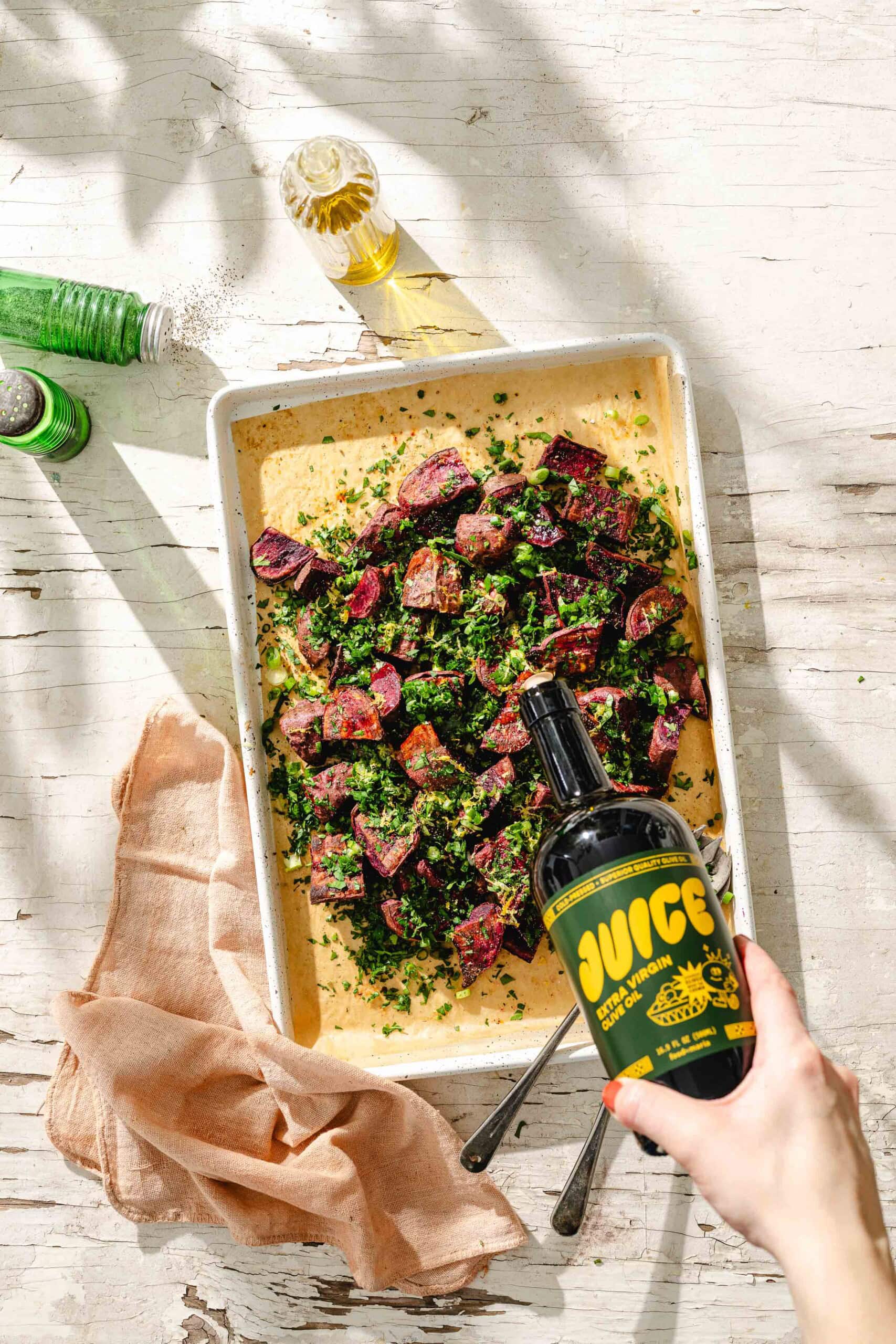 A hand holds a bottle labeled JUICE over a tray of roasted vegetables garnished with chopped herbs on a rustic white table, with a green bottle, olive oil, and a beige cloth nearby.