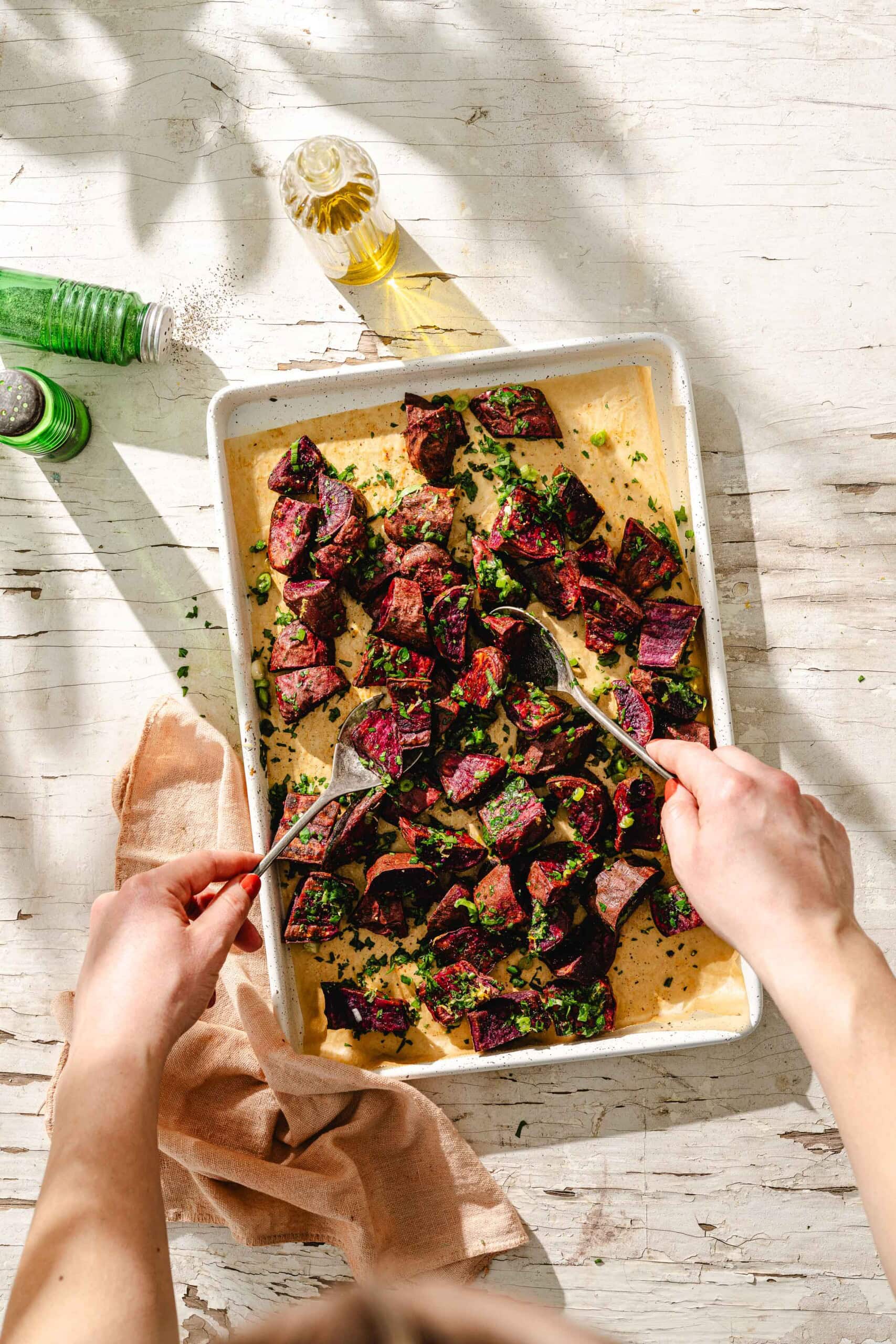 A person uses two forks to toss roasted purple potatoes garnished with herbs in a white baking dish on a rustic white wooden table, with a green bottle, oil bottle, and tan napkin nearby.