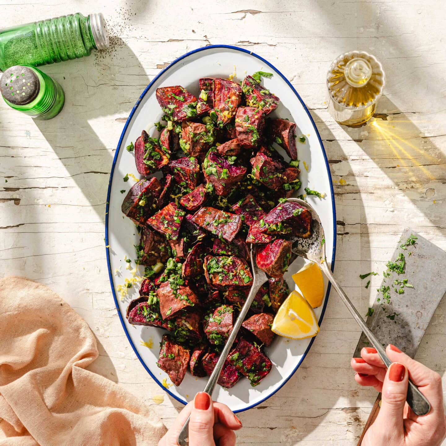 A platter of roasted beet salad garnished with chopped herbs and lemon zest sits on a white table. Two hands with red nail polish hold serving utensils and a lemon wedge. Green spice shakers and a bottle of oil are nearby.